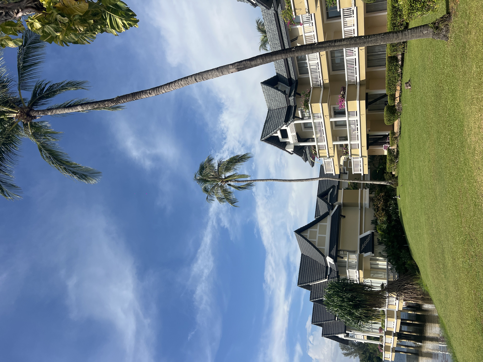The photograph shows a beachfront resort in Choeng Thale, Thailand, under a partially cloudy sky. Several palm trees are visible. A multi-story yellow building with dark-tiled roofs and white-railed balconies is present. A lawn with green grass is to the side of the building. Some of the building seems to be constructed over water. No people are visible.