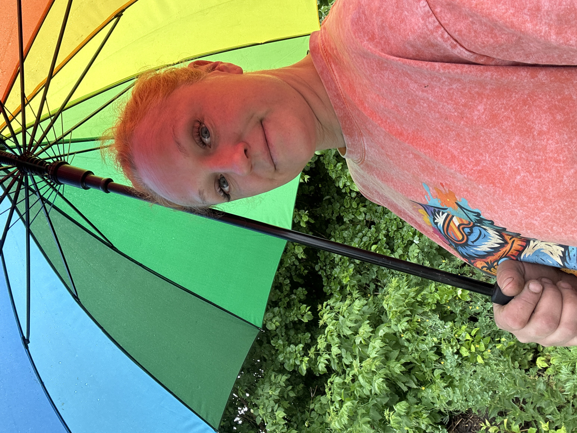 A pale-skinned woman with blue eyes is seen from a low angle, holding a colorful rainbow umbrella. She wears a pink/peach long-sleeve shirt with a design on the front. Behind her are green bushes. The photo appears to be taken outside, possibly in a garden or park-like setting.