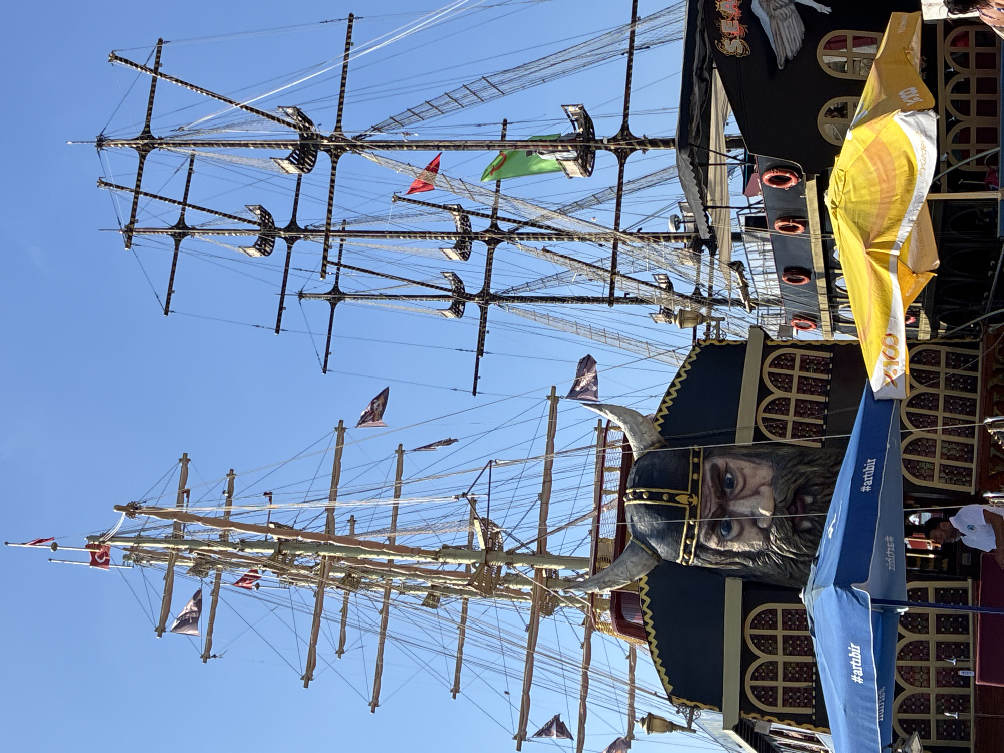 This is a close-up, eye-level, outdoor, daytime photograph of two docked pirate-themed ships. The ship on the left has a large, carved Viking face with horns on its bow. The ship on the right is black with a large white winged creature emblem and the word "SEA" visible. Both ships have tall masts with rigging and flags. In the foreground, there are two open umbrellas, one blue with the hashtag "#artibir" and the other yellow. A man wearing a white polo shirt is standing near the blue umbrella. The ships appear to be in a harbor. This scene is consistent with the popular pirate ship tours departing from the marina in Alanya, Turkey. The sky is clear and blue.