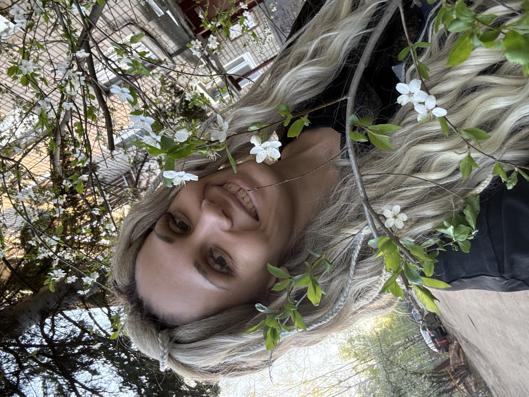 A woman with long, wavy blonde hair, featuring distinct braided sections, smiles brightly in an outdoor close-up portrait. Her face, angled upwards and partially obscured by foliage, is framed by numerous branches covered in small, delicate white five-petaled blossoms and fresh green leaves. Her eyes are open, and she looks towards the viewer, expressing a joyful or content emotion.
The setting is outdoors during bright daylight, indicative of spring. In the background, partially visible through the branches, is a multi-story brick building, suggesting an urban or suburban environment. Other trees and glimpses of the sky can also be seen. The white blossoms resemble those of fruit trees, enhancing the springtime atmosphere. The woman's dark clothing is subtly visible behind her hair.
The image captures a serene moment amidst blooming nature, located in Rusanovo, Russia. No visible text is present.