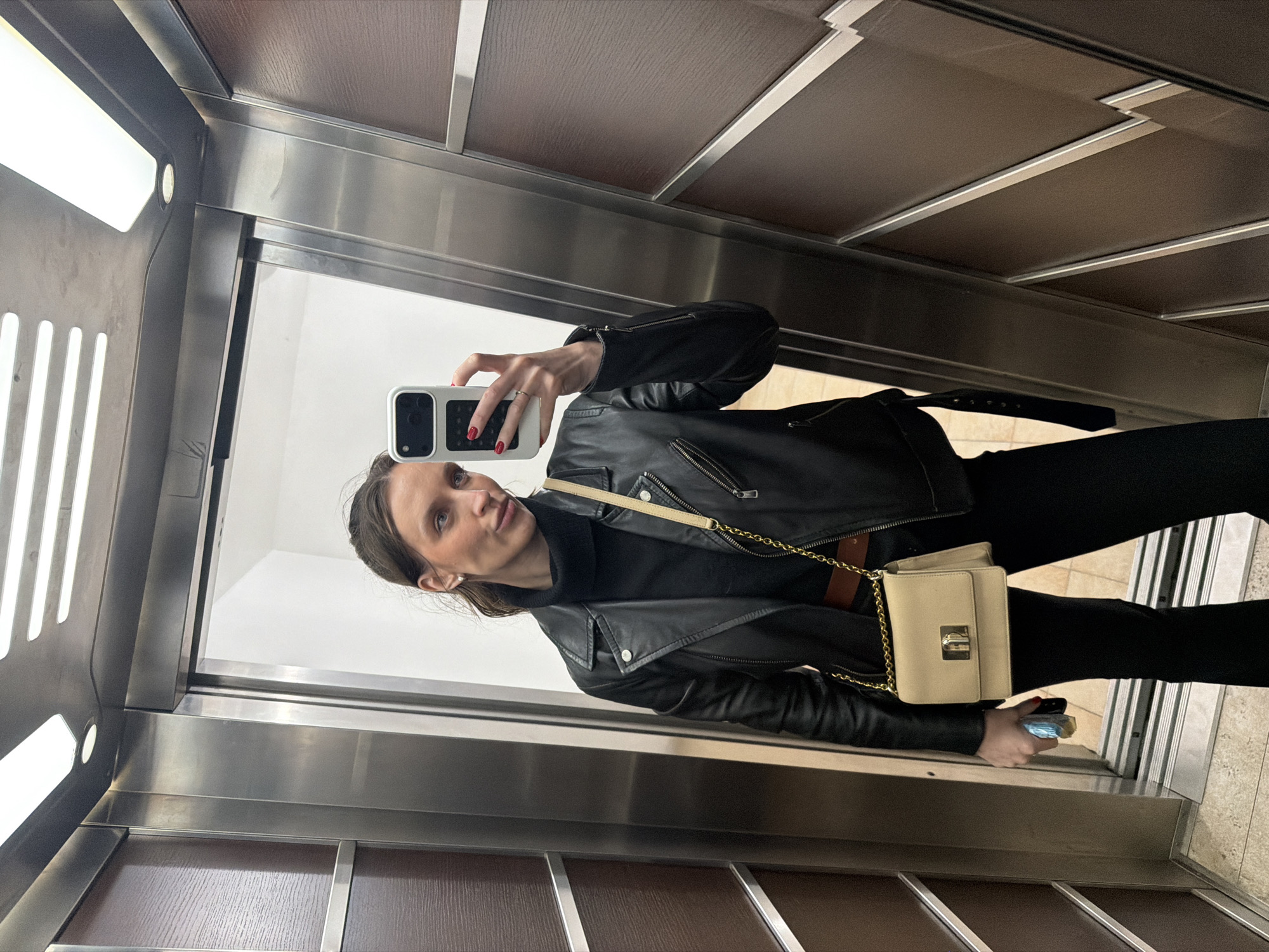 An image taken in an elevator in Budva, Montenegro, depicts a female individual capturing a mirror selfie. The subject is a young adult woman with dark hair, visible from the chest up, dressed in a black turtleneck sweater, a black leather jacket with silver-toned zippers, and black pants. A brown belt is visible around her waist. She wears a light beige shoulder bag with a gold chain strap and a gold-colored clasp, positioned across her body. Her left hand holds a white smartphone, angled to photograph her reflection, revealing red-painted fingernails and a ring on her left ring finger. Her right hand holds a small, clear blue-tinted object and a small gold-colored item. Her gaze is directed slightly to the left, with her lips subtly pursed. The elevator interior features dark brown wall panels accented with metallic horizontal and diagonal trim. The ceiling contains multiple rectangular strip lights and a circular recessed light fixture. A partially open elevator door reflects a white background. The floor is light-colored and tiled.
