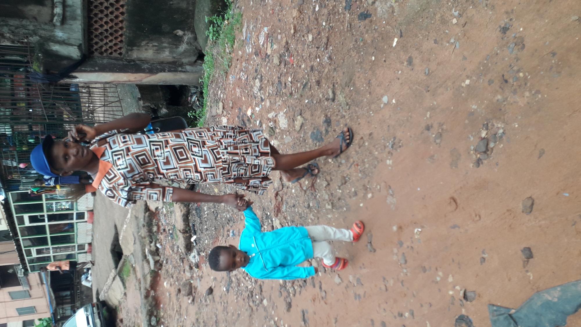 A woman is holding the hand of a young child while standing on a dirt road in what appears to be a section of Onitsha, Nigeria. The woman is wearing a brown and white patterned dress and a blue baseball cap. She is also talking on her cell phone. The child is wearing a light blue jacket, white pants, and red sandals. The road is unpaved and uneven, with various debris scattered around. In the background, buildings and structures typical of a densely populated urban area are visible. The overall impression is a snapshot of daily life in a bustling, somewhat run-down environment.