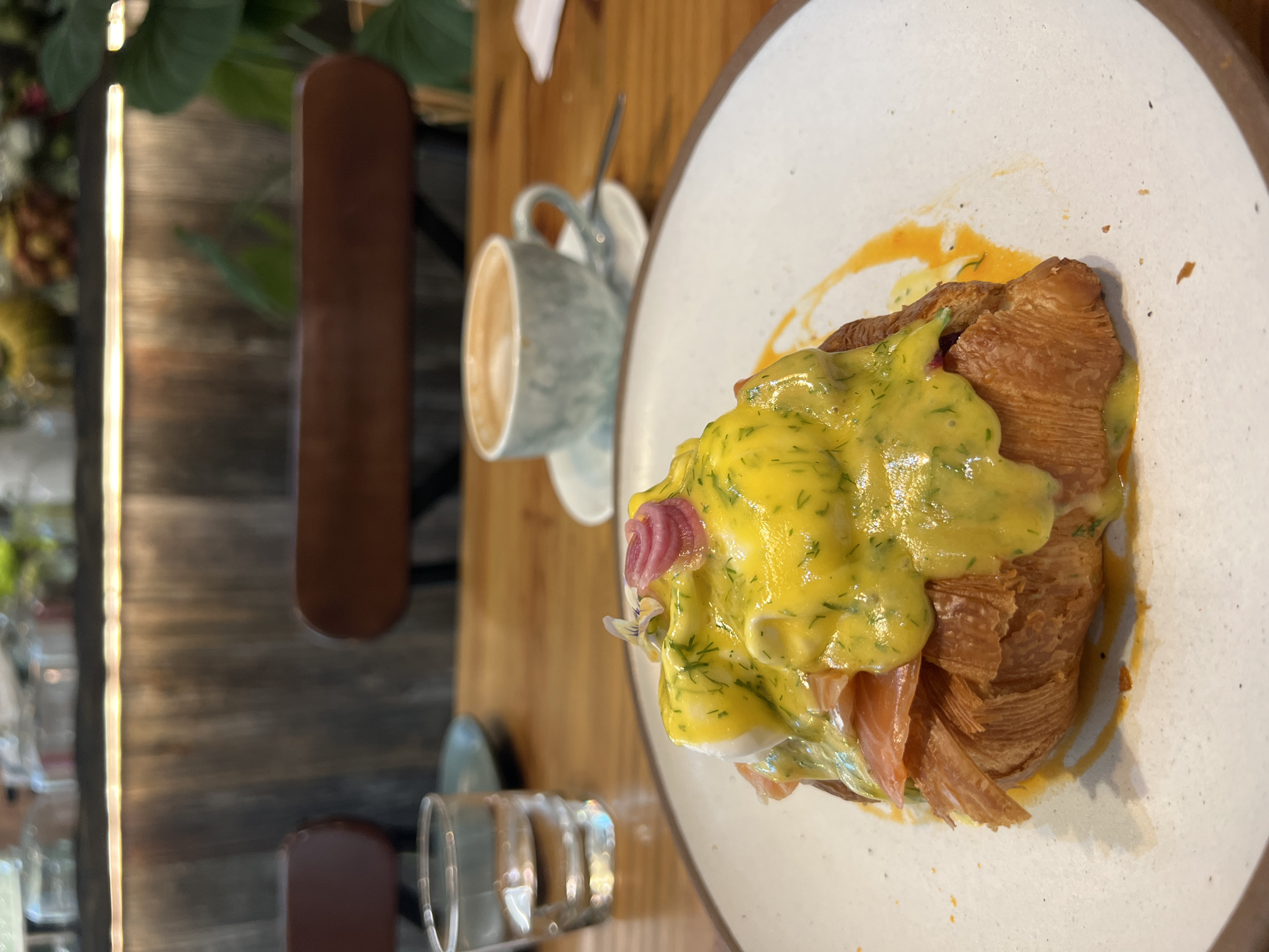 On a wooden table, a plate holds a croissant topped with salmon, a poached egg, and hollandaise sauce, garnished with dill and a pickled onion. Beside it, a cup of coffee rests on a saucer, and a glass of water is nearby. The background shows a blurred plant and chairs. The location is potentially in Nha Trang Ward, Vietnam.