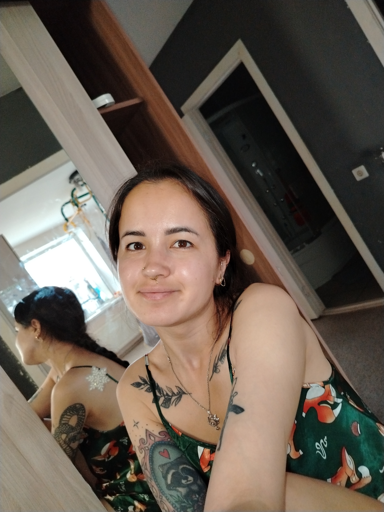 A young woman with dark hair, a nose ring, and visible tattoos on her arm and chest is taking a selfie in a room that appears to be a bedroom or living space. She is wearing a green camisole with a fox pattern. A mirror reflects her from the side, showing more of her tattoos, including a snake on her arm and a snowflake on her back. The room features wooden furniture and dark grey walls. There is a doorway in the background leading to another room. The location is identified as Noviy, Russia.