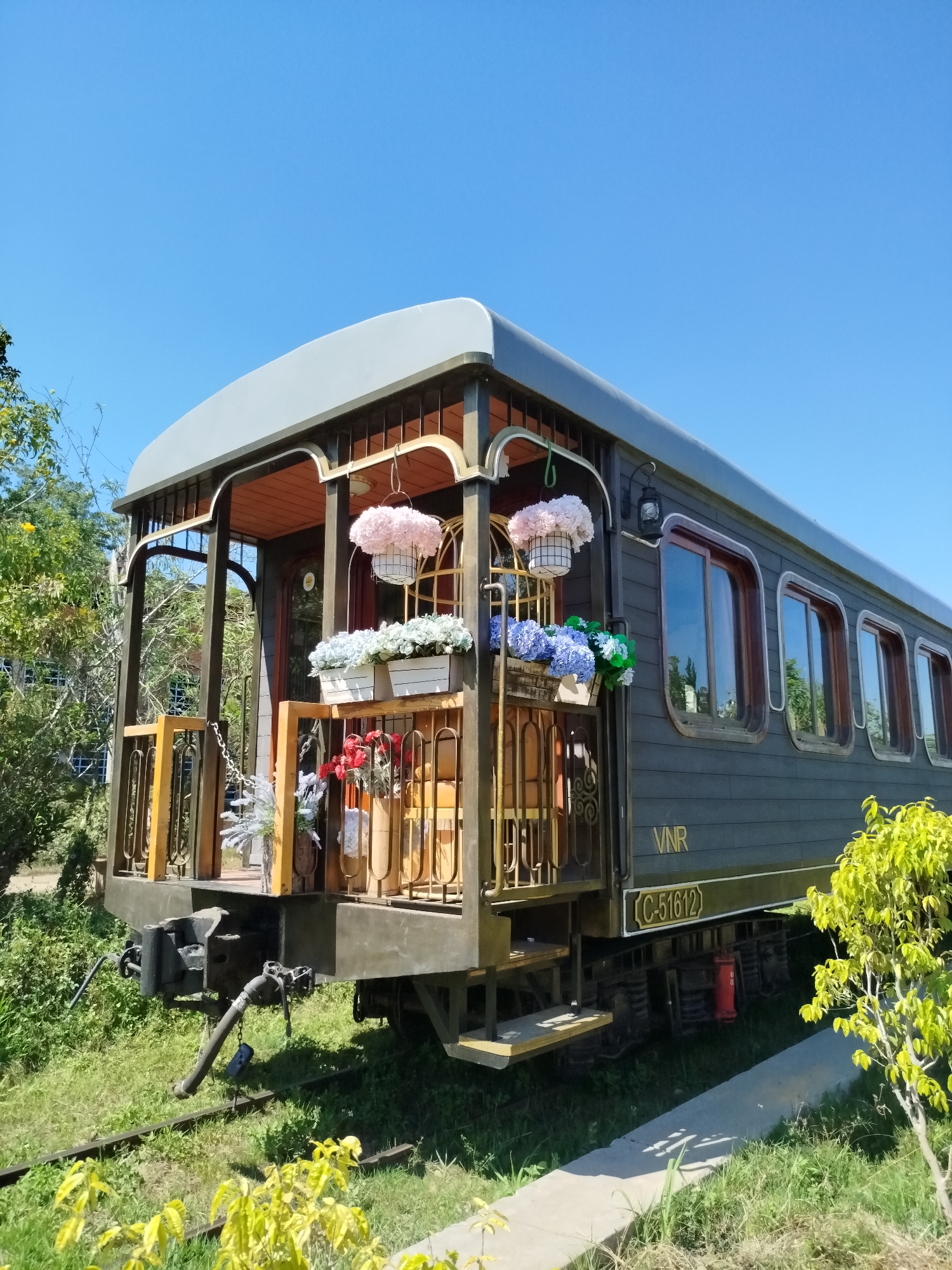 A vintage-style passenger train car, painted dark grey-green with a light grey roof, is depicted stationary on a single set of railway tracks under a clear, bright blue sky. The visible end of the car features an ornate, open-air balcony with dark metal railings and gold accents, furnished with a wooden floor. This balcony is extensively decorated with artificial flowers: a large hanging basket contains pink blooms, two rectangular planters hold white/light green and blue/green flowers, and several vases display red and white/silver decorative foliage on the balcony floor. Along the side of the car, multiple rectangular windows with rounded corners and dark wooden frames reflect the sky and surrounding greenery. Gold lettering on the side identifies the car as "VNR," and a plaque beneath reads "C-51612." The train's undercarriage, including the bogies and coupling mechanism with an air hose, is visible. The tracks are embedded in a patch of green grass and weeds, flanked by various trees and shrubs, including a small, bright green-leafed bush in the foreground. A concrete pathway runs parallel to the tracks on the right. No individuals are present. This train car is located at the Da Lat Railway Station in Thành phố Đà Lạt, Vietnam.
