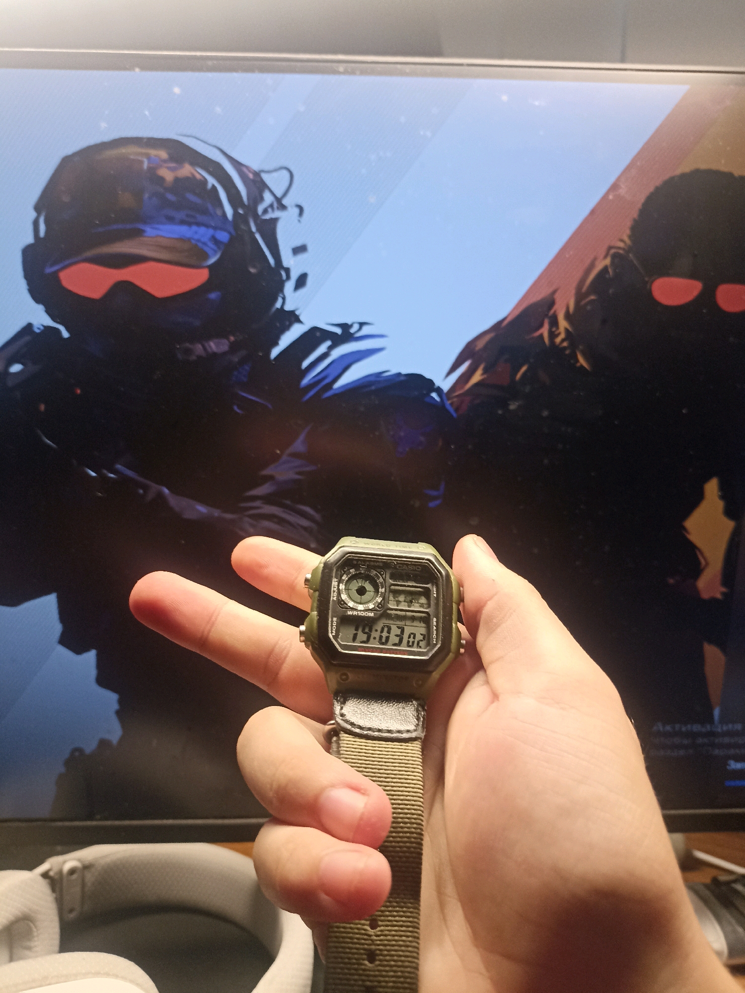 A person's hand holds a digital watch with a green face and a tan fabric strap in the foreground. The watch displays the time "19:03 02". In the background, a computer monitor displays an image of two figures wearing tactical gear and red goggles, possibly from a video game. To the right of the monitor, text in Russian, including "АКТИВАЦИЯ" (Activation), is visible, suggesting a user interface or program window. The scene appears to be indoors, likely in a home or office setting, with a portion of a pair of white headphones visible at the bottom left. There is no indication of the city of Mariupol, Ukraine, in the image.