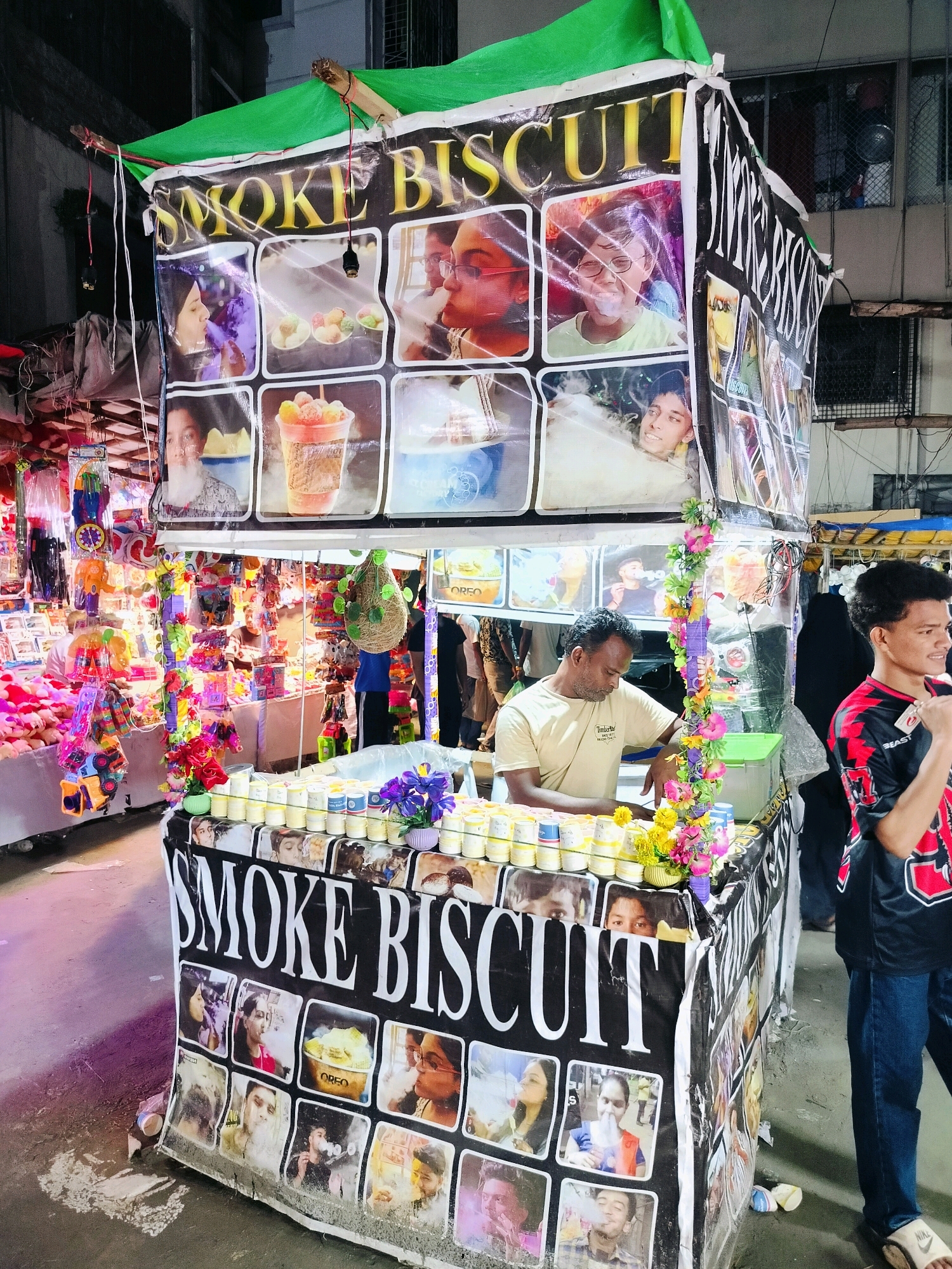 In Dhaka, Bangladesh, a street vendor operates a mobile cart adorned with a large banner displaying the text "SMOKE BISCUIT" in bold, gold lettering against a black background. The banner is further decorated with numerous small portraits of people, some of whom are shown exhaling smoke or vapor. The cart itself is filled with various colorful items, including what appear to be small containers of food or drink, along with a variety of toys and decorative elements, such as artificial flowers. Two men are visible at the cart. One man, the vendor, stands behind the counter, engaged in his work. Another man stands to the right, observing. The setting appears to be a bustling street or market, with other vendors and shoppers in the background, suggesting a lively urban environment.