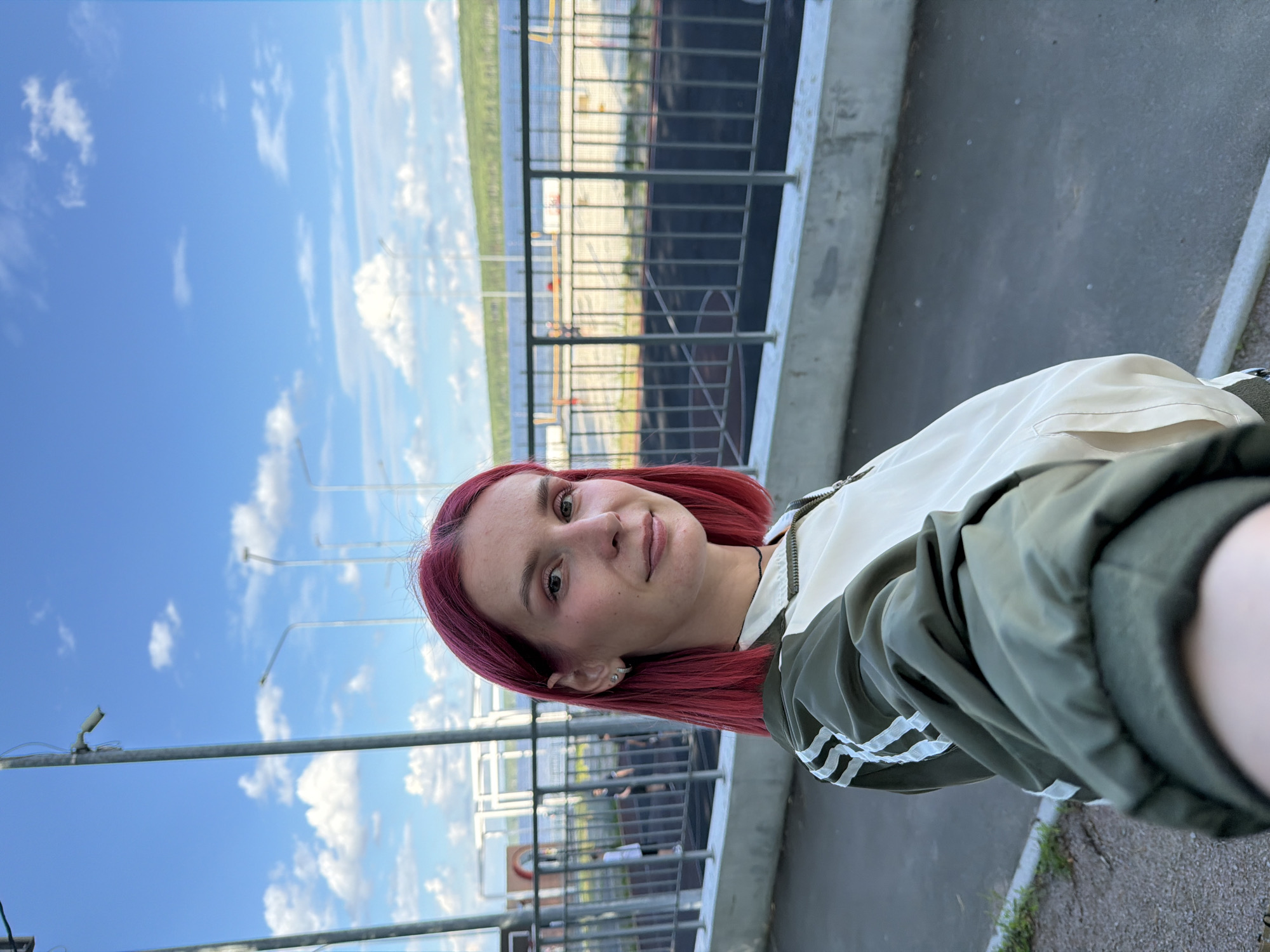 A young woman with short, vibrant red hair smiles at the camera in a selfie. She is wearing a light-colored jacket with green accents and white stripes on the sleeves. The background features a bright blue sky with scattered white clouds. A metal fence and a concrete structure are visible behind her. Beyond the fence, a body of water or a large industrial area is suggested, with some buildings and infrastructure. The overall impression is of an outdoor setting, possibly during the day with good weather. The image appears to be taken in Chaikovsky, Russia.