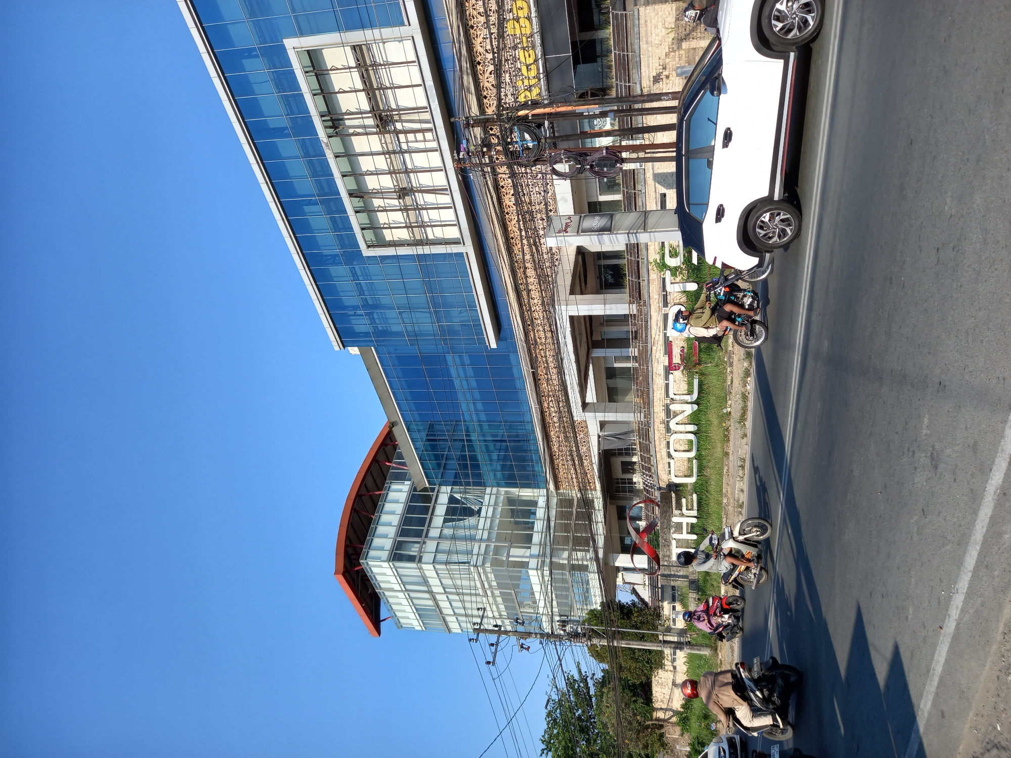 A wide shot captures a sunny day in Samarinda, Indonesia, focusing on a modern, multi-story building with a glass facade. The building, branded with large white letters spelling out "THE CONCEPT" at its base, stands against a clear blue sky. In front of the building, a street hosts traffic, with several motorcycles and a white SUV. On the street, four individuals are visible riding motorcycles. One person on a black and white motorcycle in the foreground wears a helmet and a brown jacket. Further back, another person on a red motorcycle is also visible. To the right, a white SUV with a black roof is traveling down the road, and a person is riding a motorcycle behind it. The scene is completed by power lines stretching across the frame and some vegetation on the left side of the road.