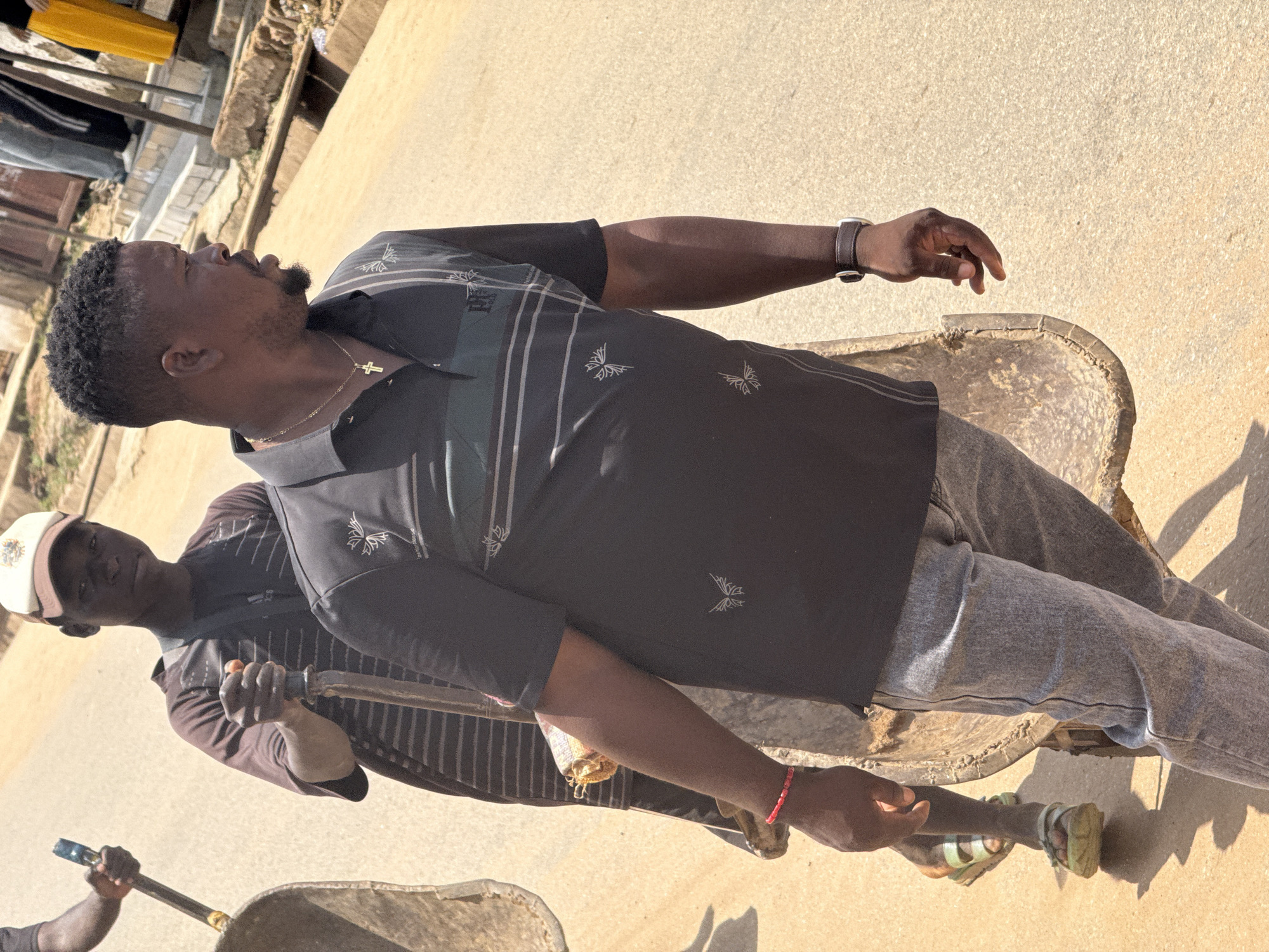 In Abuja, Nigeria, two men are seen pushing a cart laden with sand. The man in the foreground, wearing a black polo shirt with a floral pattern and grey jeans, is looking to his right. He has a watch on his left wrist and a gold chain with a cross pendant around his neck. The man behind him, wearing a striped t-shirt and a baseball cap, is holding a shovel. The cart appears to be made of rough material and is positioned on a sandy road. In the background, buildings and stairs suggest an urban environment. The lighting indicates it is daytime.