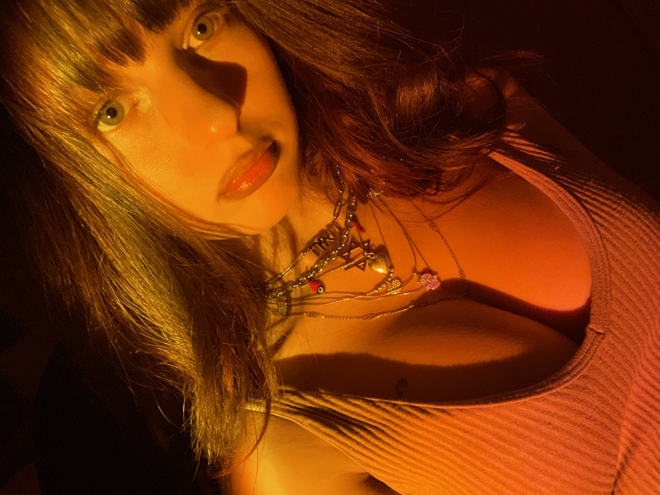 A woman gazes at the viewer with striking green eyes and glossy lips. Her hair is light brown with fringe. She wears several necklaces with charms, including a Star of David and an evil eye bead. The lighting is warm, casting a shadow across her face. She is wearing a tight ribbed tank top, the neckline of which shows cleavage.