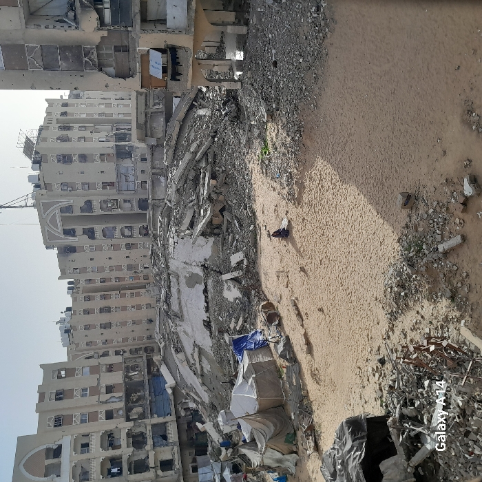 This image depicts a heavily damaged urban environment in Khan Yunis, Palestinian Territory. The foreground presents a large expanse of uneven sandy ground interspersed with concrete rubble and scattered debris. In the midground, a lone individual dressed in dark clothing is observed kneeling or sitting on the sandy surface, facing away from the camera. To the left of this individual, a cluster of makeshift shelters, constructed from white fabric and blue tarpaulins, is erected amidst the debris. Behind the shelters and the individual, a massive mound of collapsed building materials, including pulverized concrete slabs and twisted rebar, dominates the central area. The background is composed of a row of multi-story buildings, all exhibiting extensive structural damage: numerous impact holes, missing windows, shattered facades, and destroyed balconies are visible across their surfaces. Water tanks are present on the rooftops of several background buildings. The sky above is clear and light. The scene conveys a state of stillness within a landscape of widespread destruction.