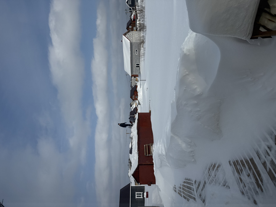 The image depicts a snow-covered residential area in Gornyi (Dvurechenskoe S/P), Russia, under a partly cloudy sky with visible blue patches. In the foreground, a wooden deck or pathway is partially exposed from under deep, undulating snowdrifts, some exceeding knee height. A dark object, possibly a table or bench, is discernible under snow on the far right. The midground features a large, rectangular, dark red metal structure, likely a shed or garage, with its roof covered in a thick layer of snow. A wooden pallet or makeshift ladder leans against its side. A single individual, dressed in dark winter clothing, stands upright on the snow-covered roof of this red structure, with a smaller, dark, indistinct object partially visible nearby on the roof. Beyond this structure, a vast expanse of undisturbed snow extends across the landscape. The background reveals multiple detached houses of various designs and colors, including a grey house with a dark roof on the far left, and a light grey, possibly unfinished, house with a red roof on the right. Utility poles and a construction crane are faintly visible in the distant background, indicating an active or developing settlement.