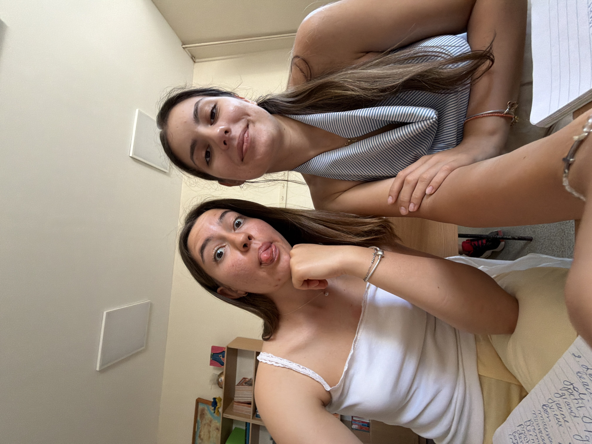 Two young women are seated at a desk, posing for a selfie. The woman on the left is wearing a white tank top and yellow shorts, with her tongue sticking out. The woman on the right is wearing a blue and white striped top. A notebook with handwritten text is visible in the foreground. In the background, a bookshelf filled with books and a world map can be seen. The ceiling features a rectangular light fixture. The scene appears to be an indoor classroom or study area, likely in Burgas, Bulgaria, given the context.