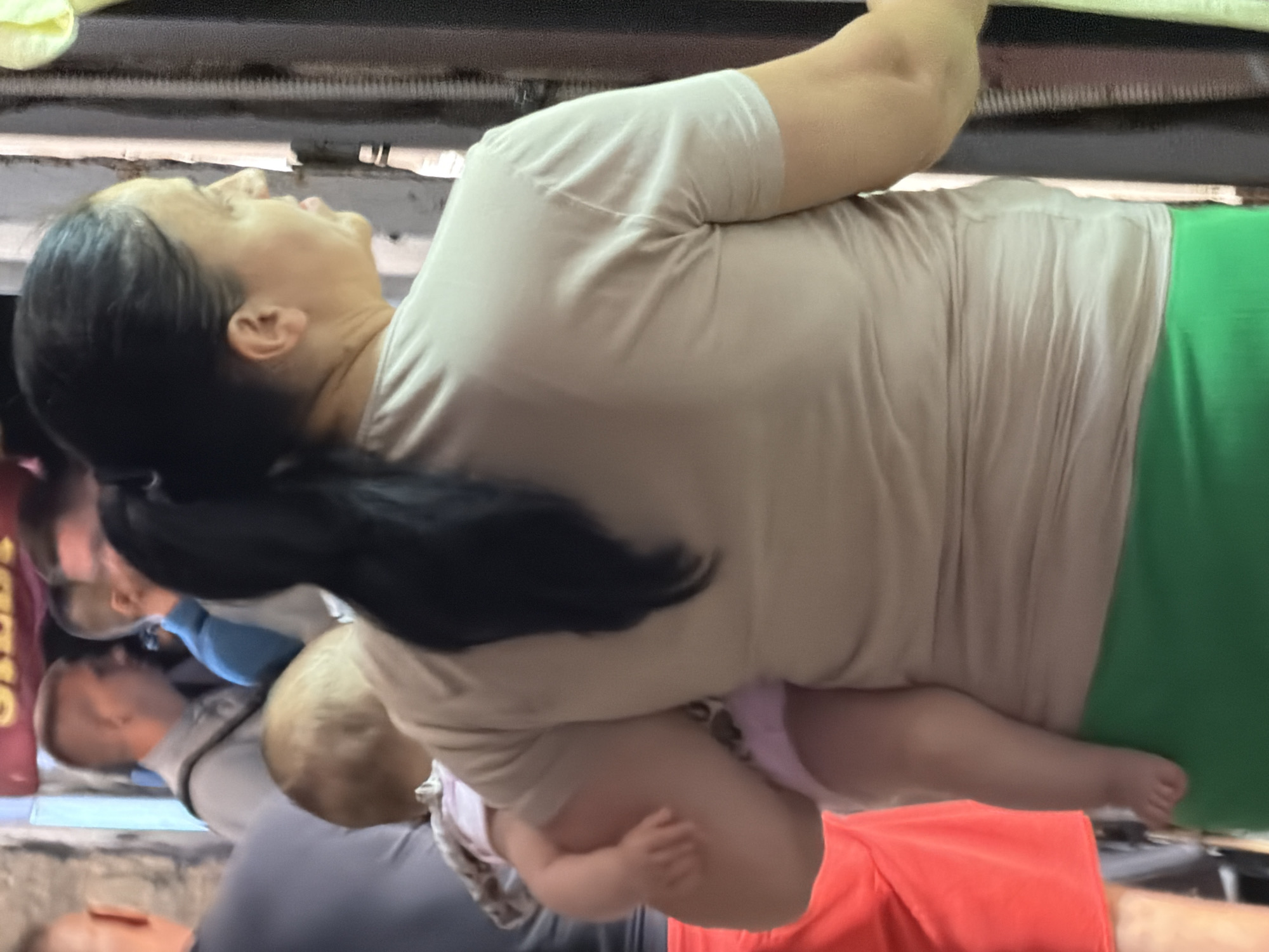 In Budva, Montenegro, a woman with dark hair pulled into a ponytail is holding a baby on her left side. She is wearing a beige t-shirt and a bright green skirt. The baby is facing away from the camera, with its left arm and leg visible. The woman is standing in an outdoor market or street, with other people visible in the background. To her left, a person wearing a red skirt is partially visible. Further in the background, several men are seen. The scene appears to be a casual outing in a public space.