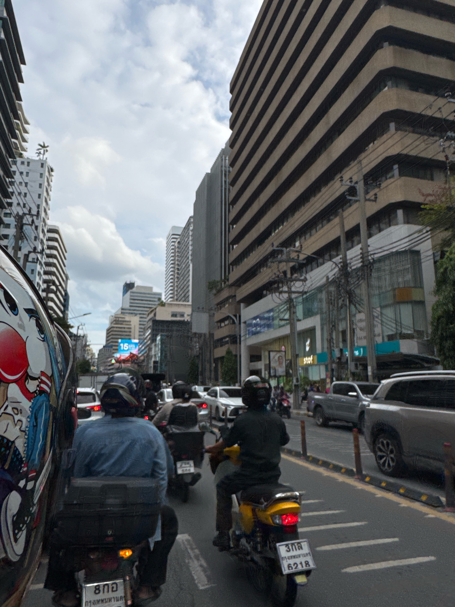 The image captures a street scene in Bangkok, Thailand, during what appears to be moderate traffic. In the foreground, a person is riding a motorcycle, their back to the camera, wearing a blue shirt and dark pants. A black storage box is mounted on the rear of the motorcycle, and a license plate with Thai characters is visible. To the left, a portion of another motorcycle's helmet, decorated with a cartoon character, is in the frame.
Several other motorcycles and cars are on the road, indicating a busy urban environment. Tall buildings line both sides of the street, characteristic of Bangkok's cityscape. One prominent building features a distinctive layered architectural design with dark horizontal bands. Another building on the right displays a glass facade with the word "stot" visible on a sign. Overhead, a network of electrical wires crisscrosses the sky, a common sight in urban Thailand. The sky is partly cloudy, suggesting daylight hours. The overall impression is a snapshot of daily life and transportation within a bustling city.