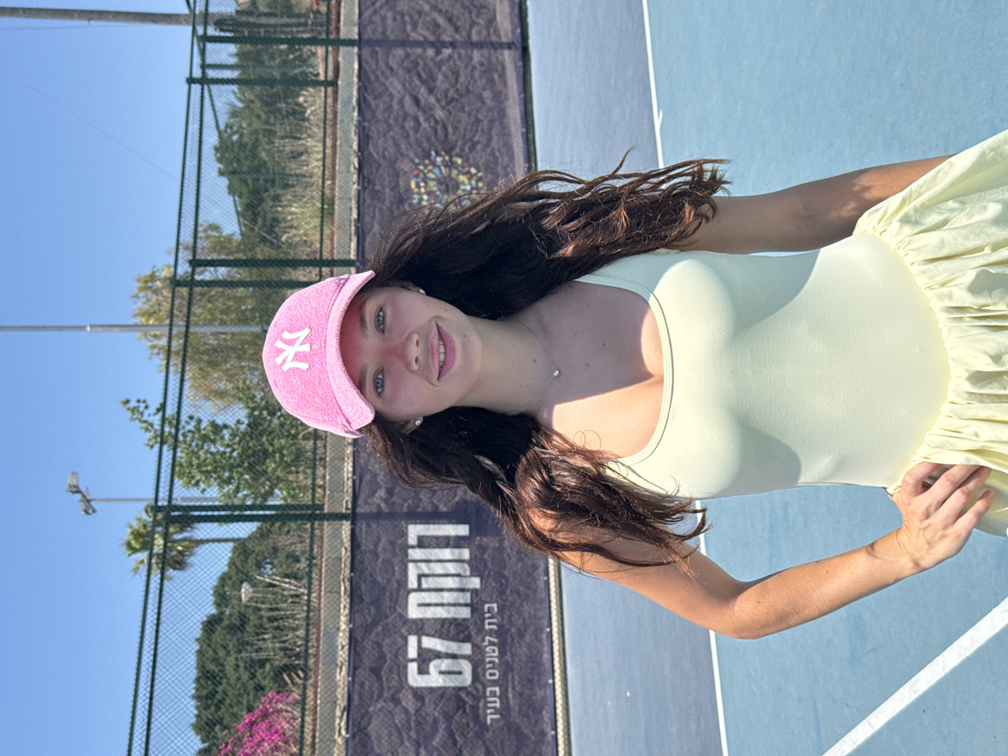 A young woman wearing a light yellow dress and a pink baseball cap with the NY logo stands on a blue tennis court. In the background, there's a fence and greenery, and behind the fence, a sign with Hebrew writing that reads "Tennis Center Beit L'Stenim Ba'Ir 67." The location is likely a tennis center in the city of Tel-Aviv, Israel, given the Hebrew writing on the sign. The woman is looking at the camera and smiling.