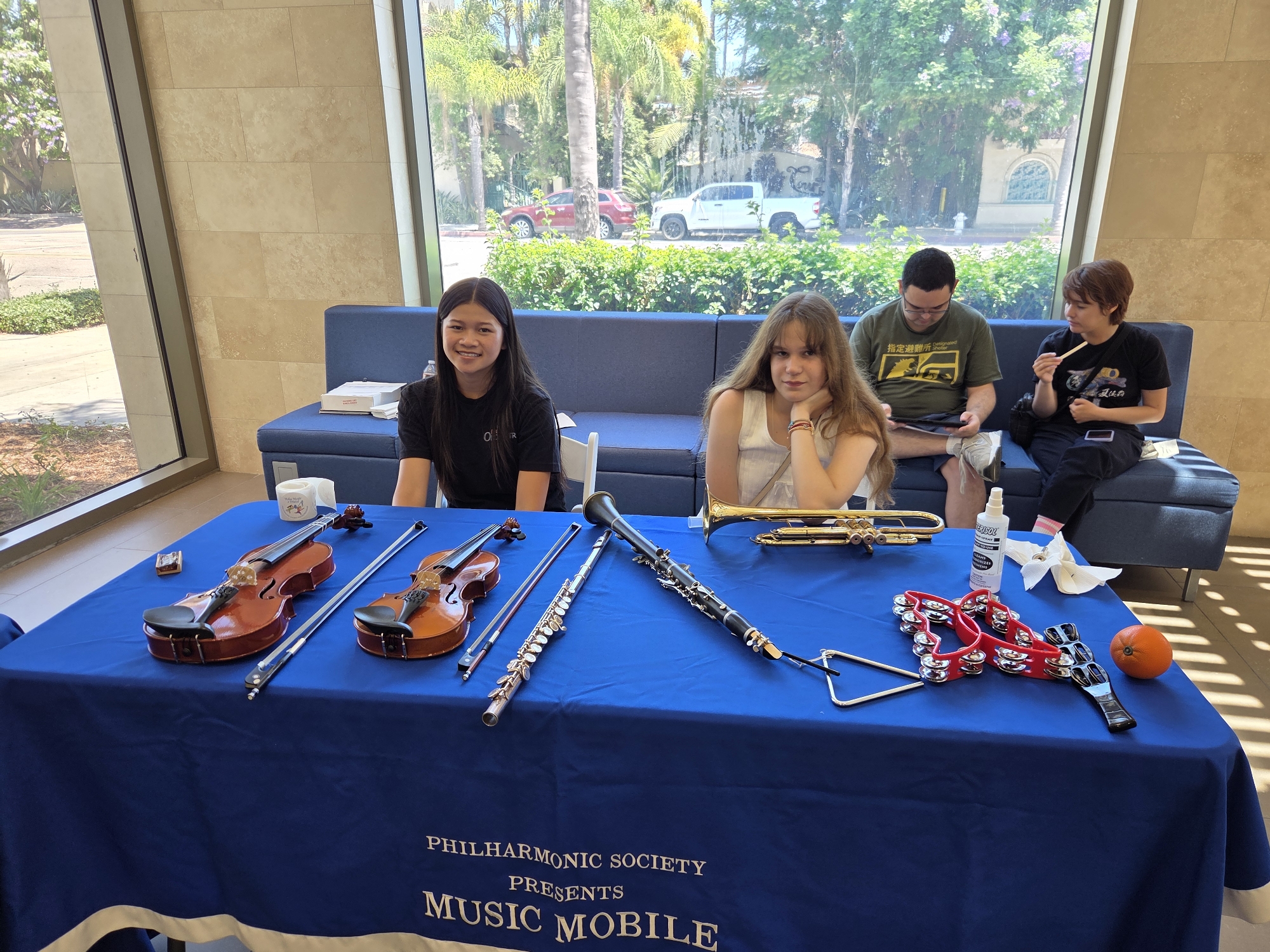In Santa Ana, California, at the Philharmonic Society's Music Mobile event, four individuals are present. Two young women are seated on a blue couch, positioned behind a table draped with a blue tablecloth. The table displays various musical instruments, including two violins, a clarinet, a flute, a trumpet, a triangle, and a tambourine. An orange sits at the end of the table. A bottle of hand sanitizer is also visible. In the background, a large window offers a view of palm trees and vehicles on a street. One young woman on the left is wearing a black t-shirt and smiling. The young woman to her right is wearing a white top and resting her chin on her hand. Further down the couch, two individuals are seated, one in a green t-shirt, and another in a black t-shirt. The environment appears to be indoors, likely a public space with a modern architectural design featuring beige tiled walls.