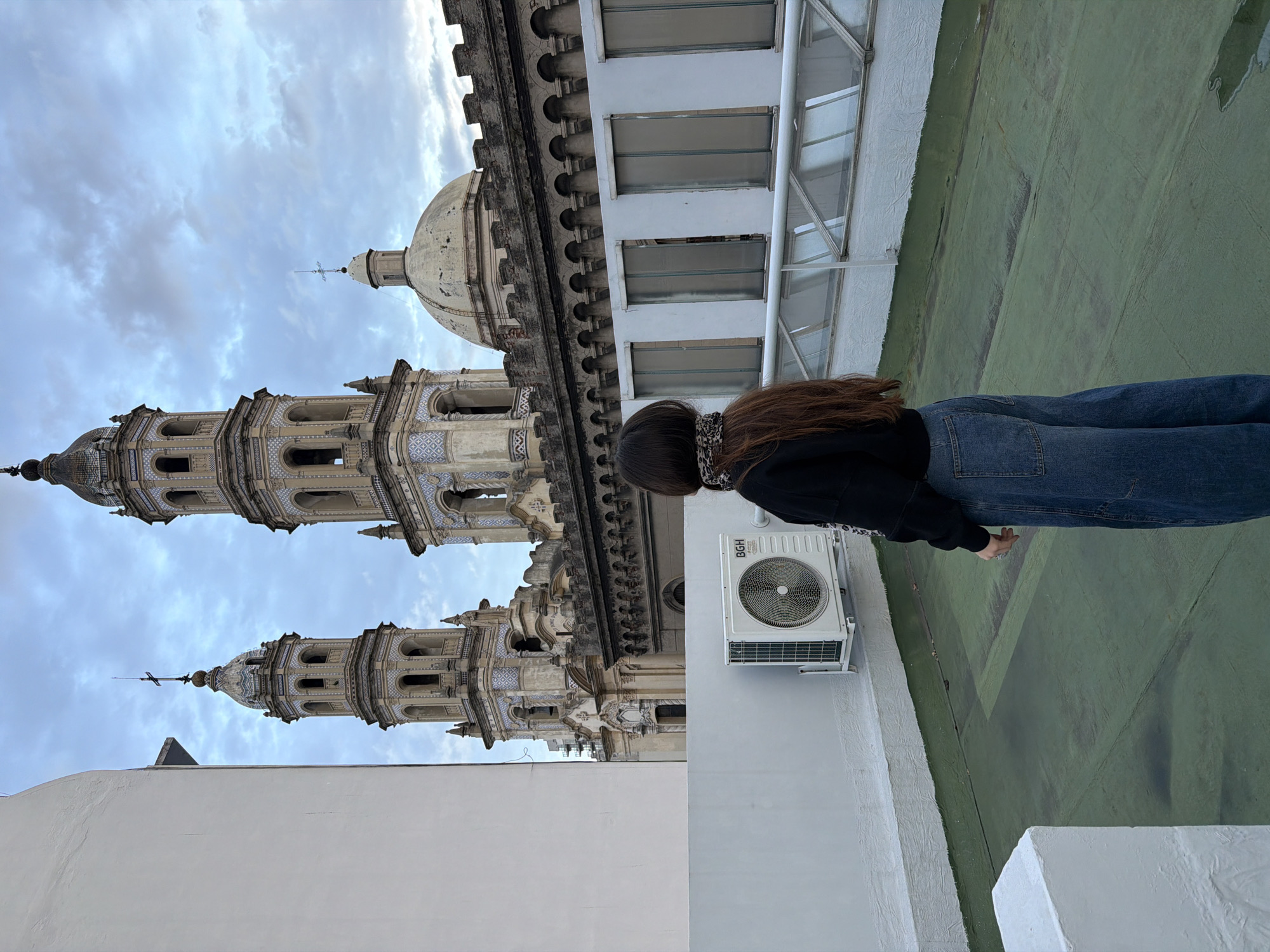 A medium shot, taken at an extreme oblique angle, shows a person standing on a rooftop in Buenos Aires, Argentina. The subject, seen from the mid-back, has long, dark brown hair and is wearing a black sweatshirt, blue jeans, and a patterned neck covering. The person is standing near the edge of the building, with a section of a green-painted rooftop visible. An air conditioning unit is attached to a white wall behind the person. In the background, there is a partial view of a building with white walls and multiple windows, and beyond that, the Basilica of Our Lady of the Pillar, a distinct baroque church known for its tiled domes and twin towers. The sky is overcast.