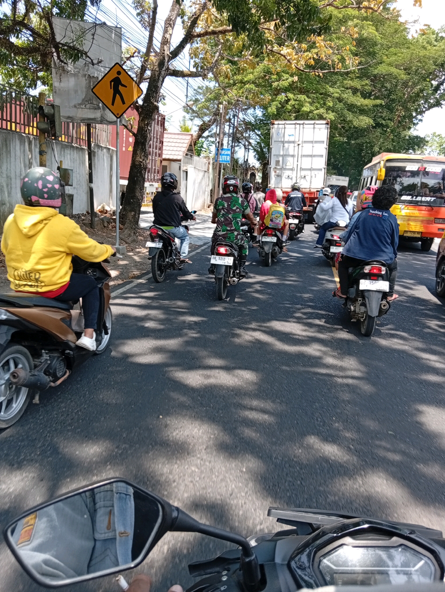 This image captures a traffic scene in Watutumou Tiga, Indonesia, likely on a sunny day given the shadows. Several motorcycles are moving in the same direction on a paved road. In the foreground, a person is riding a brown motorcycle, wearing a yellow hooded jacket and a helmet with a heart pattern. Their reflection in the side mirror shows them wearing denim pants. Behind them, other motorcyclists are visible, some wearing helmets and casual clothing.
The environment includes trees lining the road, casting dappled shadows on the asphalt. A pedestrian crossing sign is prominently displayed on the left side of the road, near a wall and some vegetation. Further down the road, a large white shipping container is being transported by a truck, and a yellow and orange bus is also present in the traffic flow. The road appears to be an urban or semi-urban setting with buildings visible in the background. The overall impression is a snapshot of everyday traffic in an Indonesian city.