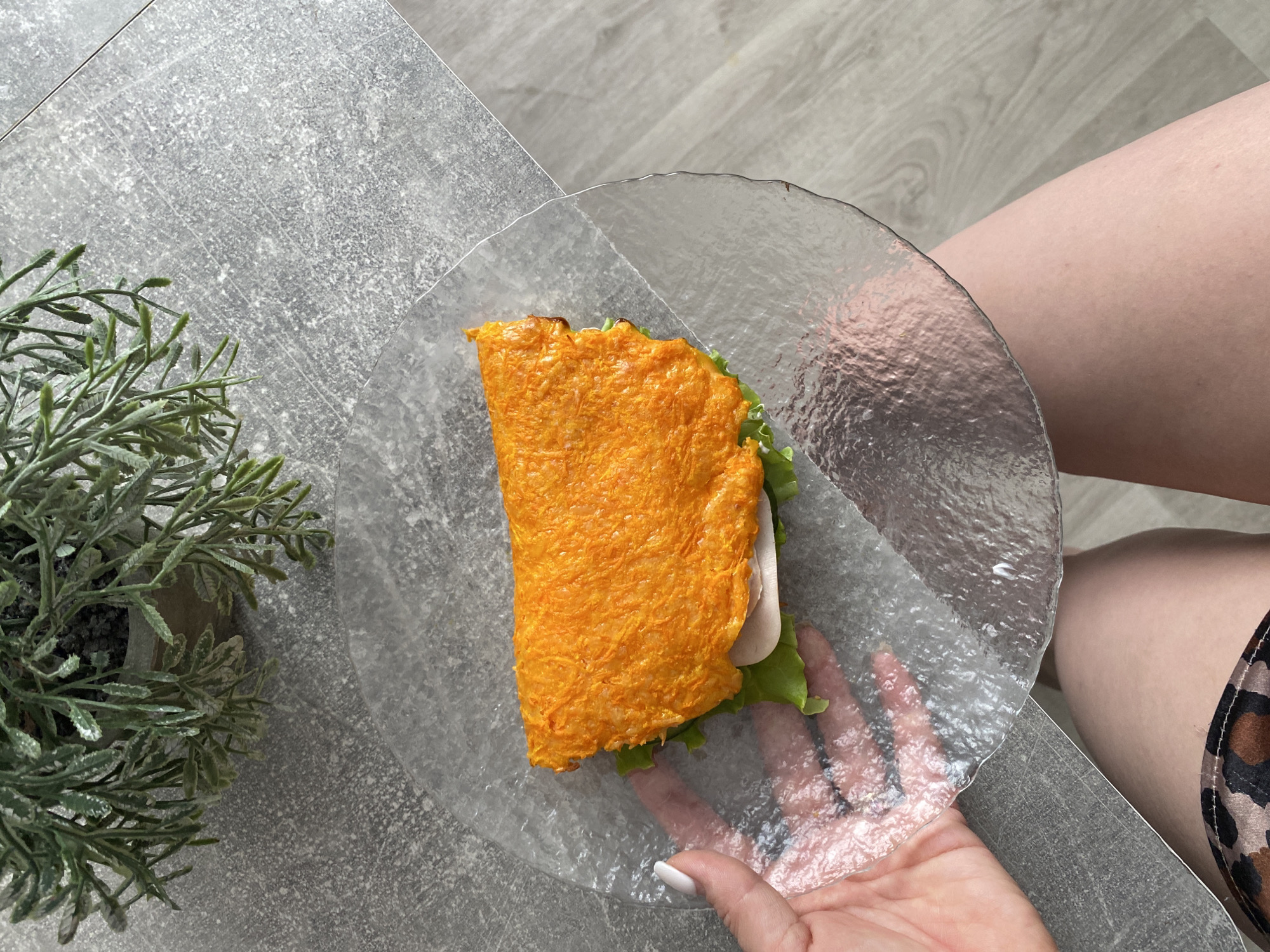 The image depicts an overhead view of a semi-circular sandwich on a clear, textured glass plate, held by a person's hand with white nail polish. The sandwich consists of an orange-colored outer layer, possibly made of cheese or carrot, with visible lettuce and a slice of what appears to be white meat (turkey or chicken). The plate rests partially on a gray surface with a textured pattern, possibly a table or countertop, and is also near a potted green plant with delicate leaves. Part of a person's leg is visible to the right, showing pale skin and clothing with a brown and black animal print pattern. No specific location in Chaikovsky, Russia, can be determined from the image.