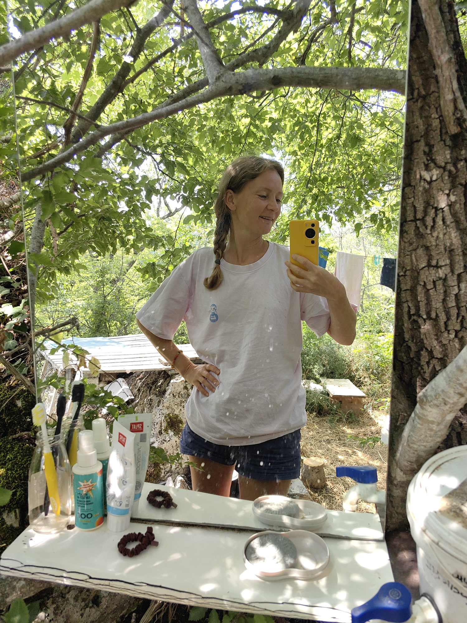 A woman with blonde hair braided to the side takes a mirror selfie outdoors. She is wearing a light pink t-shirt and denim shorts. She holds a yellow smartphone in her right hand. The mirror reflects a scene of lush greenery, with trees and leaves overhead. In the background, laundry hangs on a clothesline. In the foreground, a white shelf holds toiletries such as toothbrushes, sunscreen, and toothpaste, as well as two hair ties. A faucet with a blue handle is visible in the reflection. The setting appears to be rural, potentially a campsite or a backyard. The image does not provide enough information to pinpoint the specific location in Utios, Ukraine.