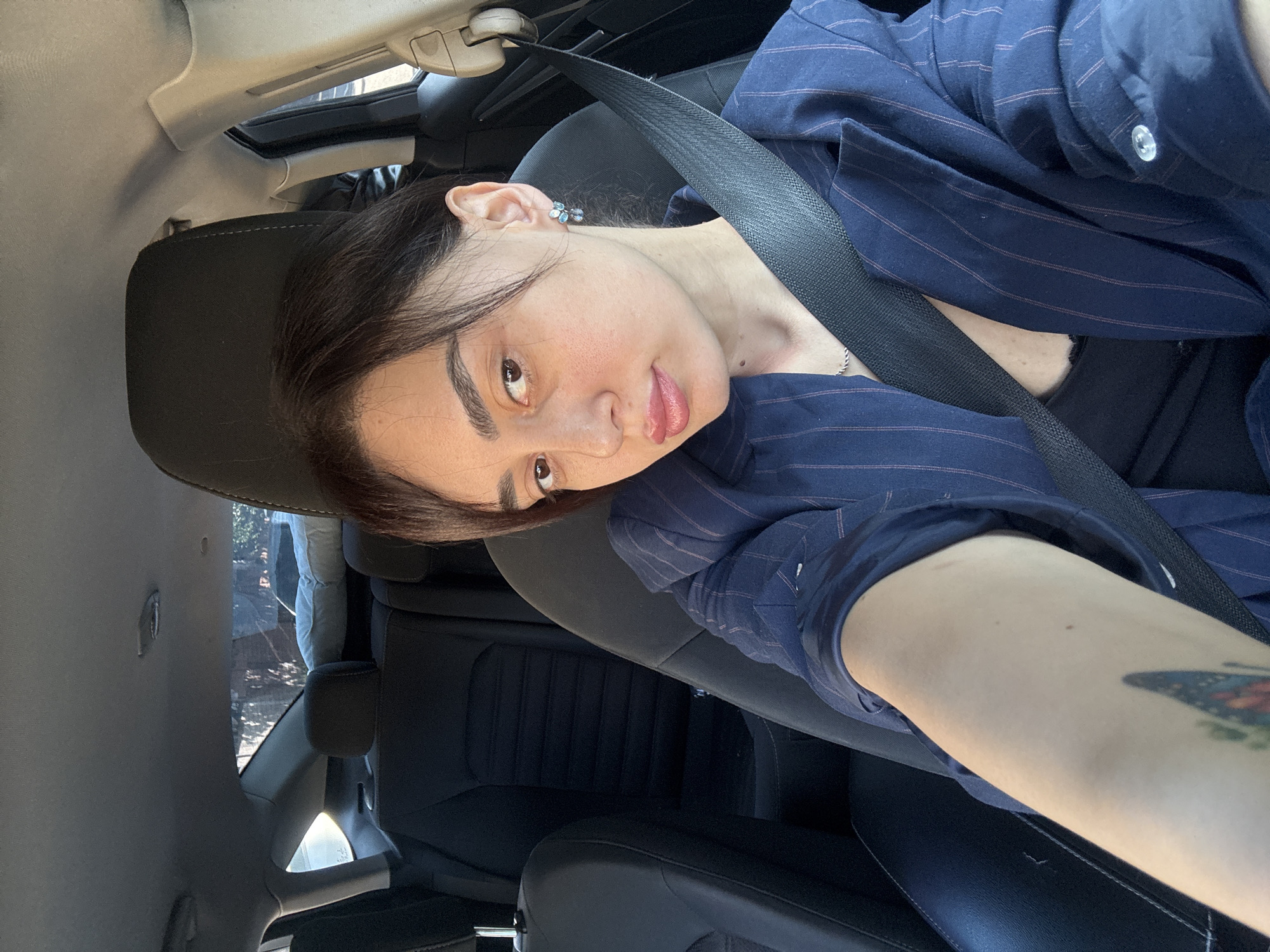 A woman is in a car. She is wearing a blue pinstripe shirt over a black top and a seatbelt. She has dark hair and light skin and is wearing earrings. A butterfly tattoo is visible on her forearm. The car's interior is black. The setting appears to be a sunny day. It is not possible to determine the city or country from this image alone.