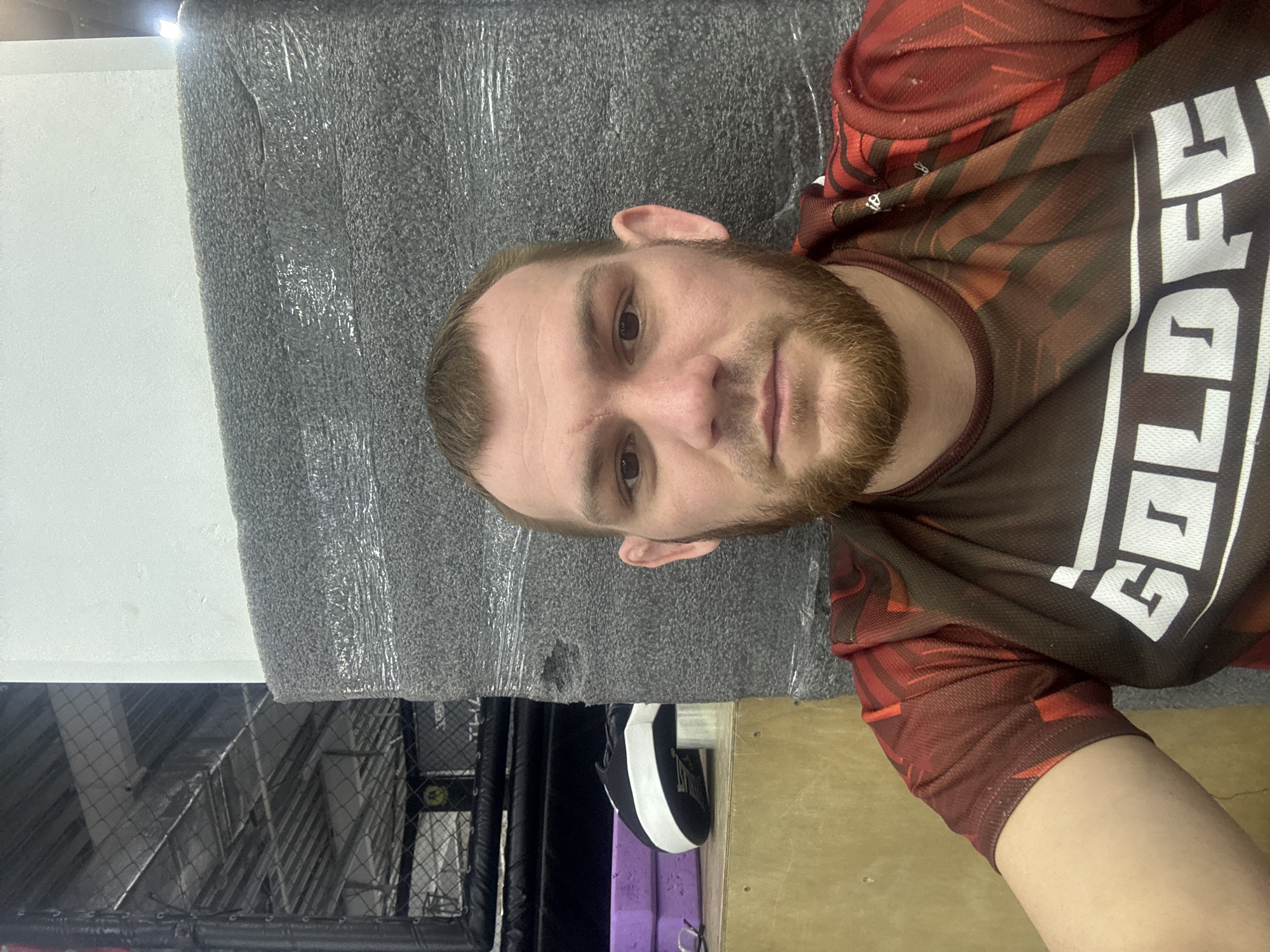 This media file shows a close-up image of a man, rotated 90 degrees clockwise. The setting appears to be a combat sports gym or training facility in Perm, Russia.
The central subject is a man with a light complexion, featuring a short, light brown beard and mustache. His hair is also light brown and closely cropped. A distinct scar or mark is visible on his forehead, above his left eye (viewer's right). He is looking directly into the camera with a neutral expression, suggesting he is taking a selfie or posing for a photo.
He is wearing a short-sleeved athletic jersey or rashguard. The shirt has a dynamic pattern of red, black, and brown geometric shapes and lines. Prominent white block letters are printed on the front, partially visible, spelling "GOLDE," likely part of a team or brand name such as "GOLDEN."
The man's head and upper body are resting against a dark grey, textured padded surface, which is covered in clear plastic film, possibly indicating protective wrapping for new or clean equipment. This padding appears to be a wall pad or a mat.
In the background, visible beneath the padded surface (relative to the image's rotation), elements of a gym are discernible. A metal mesh fence or cage, typical of an MMA or boxing facility, is clearly visible. Behind the fence, various structural elements, possibly metal beams or interior architecture, can be seen. To the lower right, a wooden or light-colored floor is visible. To the upper left of the padding, a plain white or light grey wall forms part of the backdrop.
Additional objects include a black and white boxing mitt or focus pad and a purple object (possibly another piece of gym equipment or padding) situated beneath the padded surface. The man appears to be resting or taking a break from training, given the athletic attire and gym equipment.