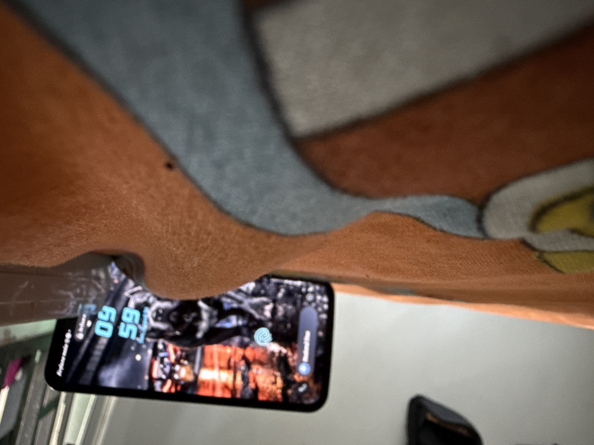 An extreme low-angle, eye-level shot captures a smartphone lying face up on a textured orange blanket with a gray patterned border. The phone, positioned horizontally, displays a lock screen with a dynamic wallpaper featuring a dark, fiery scene. The time is displayed in bright blue digits: 59:59. To the left of the time, a translucent blue notification bubble with a white dot is visible, and a circular light blue Wi-Fi symbol is present to the right of the time. The blanket appears to be a bedspread or similar fabric. The background is a plain, light-colored wall, with a blurred dark object in the upper left corner, possibly a piece of furniture or a clothing item. The overall lighting suggests an indoor environment, likely at night or in a dimly lit room. The scene is set in the city of Uyo, Nigeria.