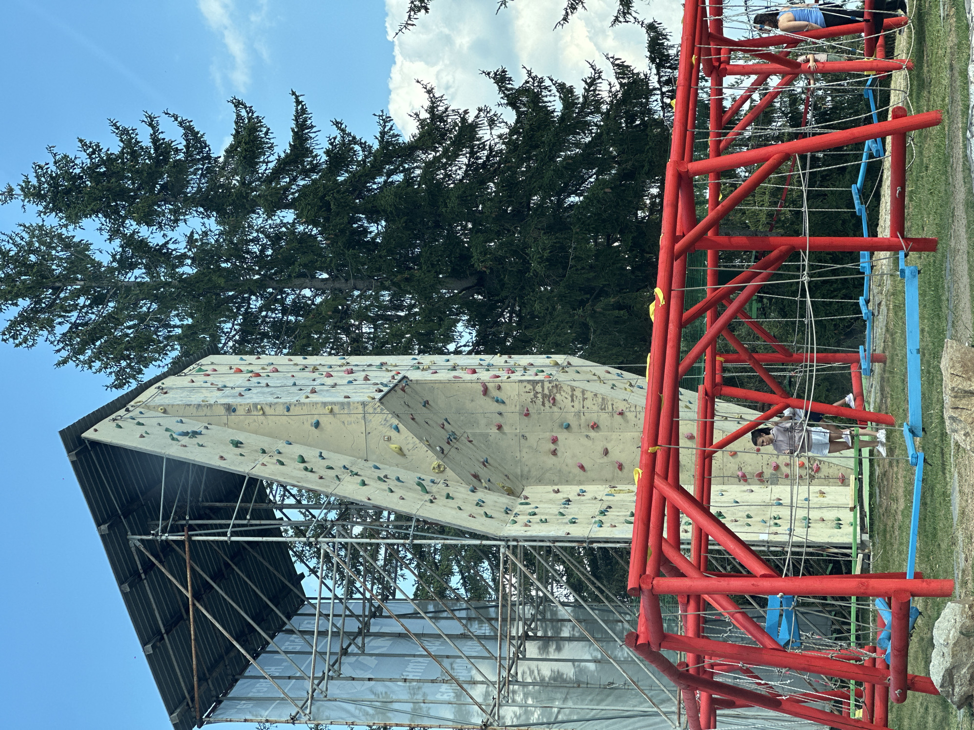 The image shows an outdoor adventure park facility in Smolyan, Bulgaria, featuring a tall, artificial climbing wall made of pale material with colorful climbing holds. To the left of the climbing wall, a scaffolding structure is visible, partially covered by a translucent tarp. In the foreground, a bright red wooden obstacle course structure with ropes and blue platforms is present. Two individuals are visible on the obstacle course. One person, wearing a light-colored hoodie and shorts, stands near the climbing wall. Another person, wearing a blue tank top and dark pants, is on the right side of the obstacle course. The background consists of lush green trees, a bright blue sky, and scattered white clouds.