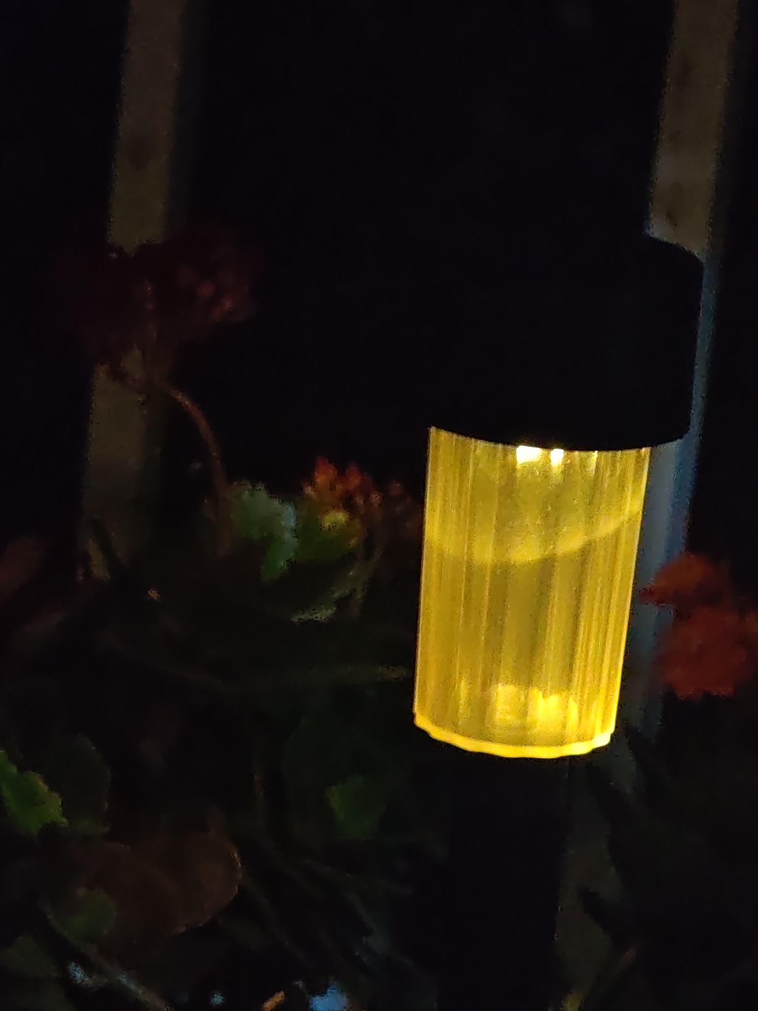The image depicts a close-up, low-light view of a solar-powered garden light illuminated at night in the city of Shumen, Bulgaria. The central object is a cylindrical light fixture, emitting a bright, warm yellow glow through its vertically ribbed, translucent body, topped by a flat, circular black cap. The intense yellow light creates a high contrast with the predominantly dark background. In the soft-focus periphery, indistinct dark green foliage and clusters of reddish plant matter, possibly flowers or berries, are visible on both sides of the light. The overall environment is outdoor and nocturnal, with the artificial light source being the primary illuminant. No individuals or specific actions are discernible within the frame.