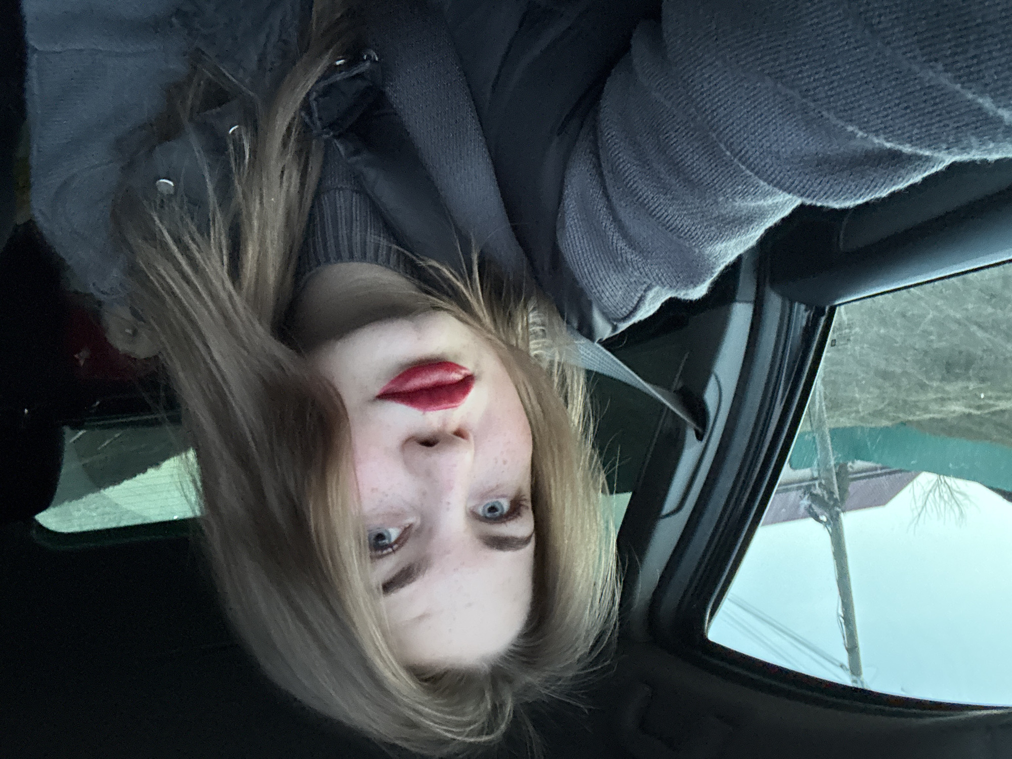 A young woman with fair skin, light brown hair, and blue eyes is seated in a car, wearing a gray sweater and a seatbelt. She has red lipstick on. The car is moving through a rural area with a few houses and a utility pole visible through the window. The location is likely in or near the city of Olkhovka, Russia.