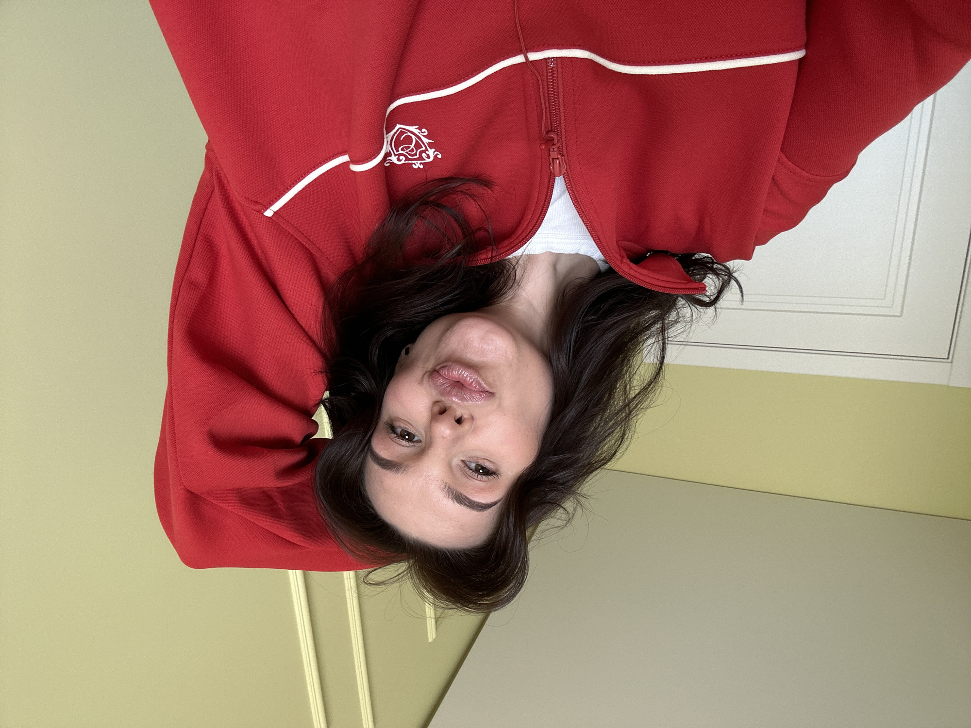 A woman with dark hair, wearing a red zip-up jacket with white trim, is looking directly at the camera. The image is rotated upside down. The background consists of a light yellow wall and a white door with decorative molding. The image was taken in Chaikovsky, Russia.