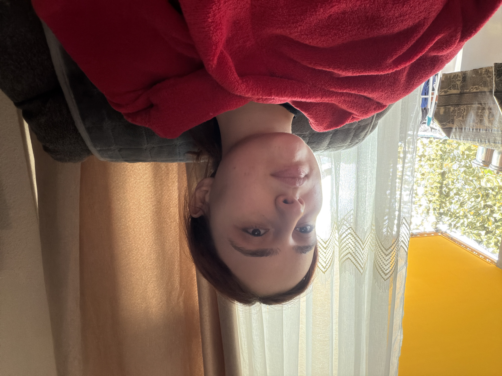The original image is rotated 90 degrees clockwise, depicting a young woman upside down, likely a selfie-style portrait taken from a low angle. Describing the scene as if correctly oriented:
The main subject is a young woman, seen from the chest up, with fair skin and dark brown hair. She looks directly at the camera with a neutral, slightly pensive expression. She has some minor blemishes on her face. Her upper body is covered by a soft, fuzzy red garment or blanket, which appears to be a bathrobe or similar loungewear. She seems to be resting or reclining, possibly on a dark grey, textured piece of furniture, like a sofa or bed headrest.
Behind the woman, to her left, is a plain light brown or beige curtain. To her right, a sheer white curtain with a distinctive gold or yellow zigzag pattern allows natural light to filter through. Beyond this window, bright sunlight illuminates vibrant green foliage from trees or bushes, and a distinct yellow wall or structure is visible. In the upper right corner, a patterned fabric (possibly a towel) and some metallic objects are partially visible, suggesting an outdoor or balcony area beyond the window.
The scene is indoors, likely a room with a window, during daytime, indicated by the bright natural light. The weather appears sunny. There are no specific visual cues within the image itself to confirm the provided location context of Strasbourg, France. No visible text is present in the image.