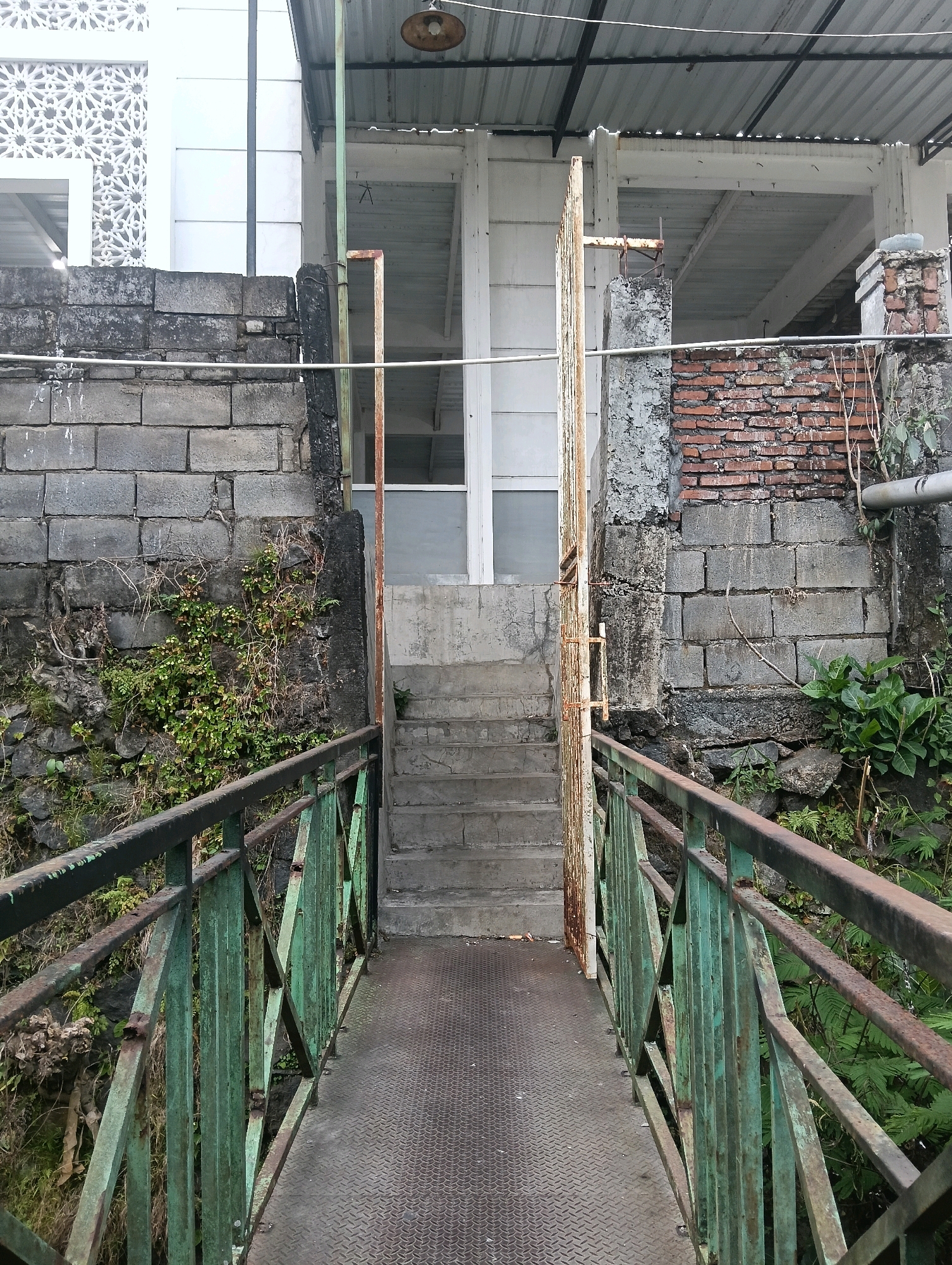 The image depicts a rusty green metal bridge with a diamond plate walkway, leading towards a set of concrete stairs. The stairs ascend to a building with a white facade featuring decorative geometric patterns and a white door. Adjacent to the stairs is an open, weathered metal gate. The bridge and stairs are flanked by stone and brick walls covered in vegetation. The building appears to be part of a complex with an overhanging corrugated metal roof and exposed beams. The scene is set outdoors, suggesting an urban or semi-urban environment in Tanjungtirto, Indonesia. No people are visible in the image. The overall impression is one of age and disrepair, with a focus on architectural elements and the bridge structure.