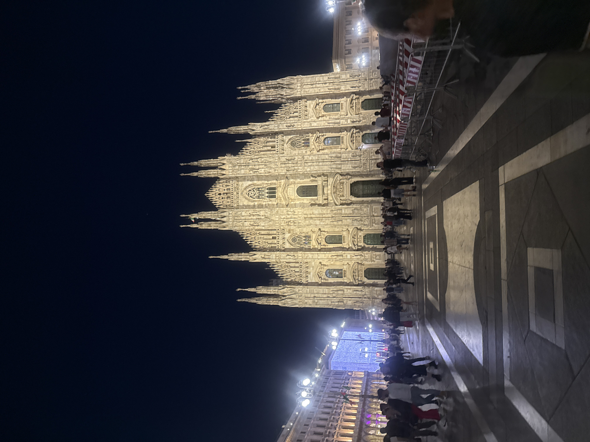 At night, a vertical shot captures the Milan Cathedral illuminated, with a crowd gathered in front of it. The building is brightly lit, contrasting with the dark sky. Pedestrians are milling around, with some close to metal barriers on the right. The foreground features a patterned stone plaza, and buildings with bright lights can be seen in the distance.