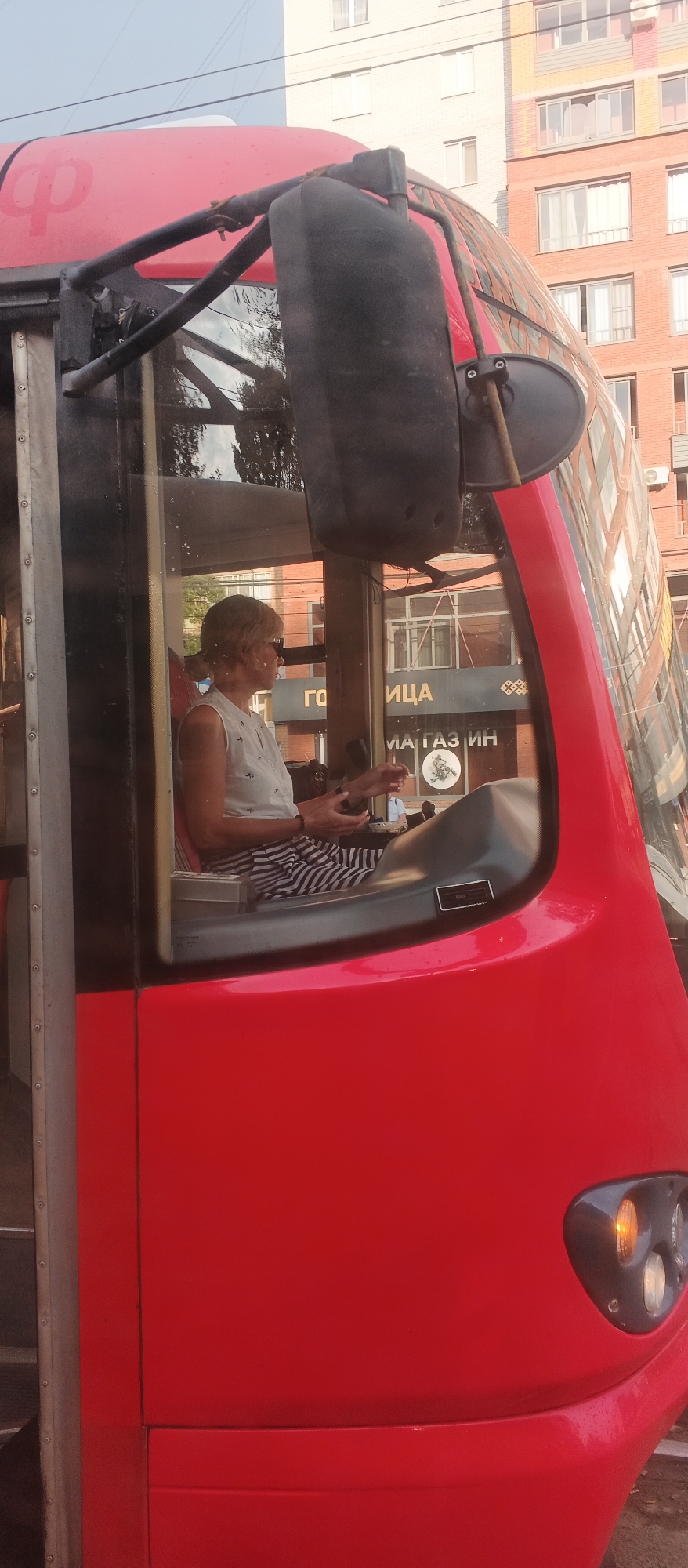 A woman with blonde hair and sunglasses is sitting in the driver's seat of a red tram. She is wearing a white sleeveless shirt with a pattern and black and white striped pants. Her hands are resting on the steering wheel and control panel. Through the window, a building with several stories and windows is visible, along with trees. The tram is likely in a city as indicated by the buildings. The logo on the tram appears to be the Russian Railways logo. This is a street-level shot taken during the day.