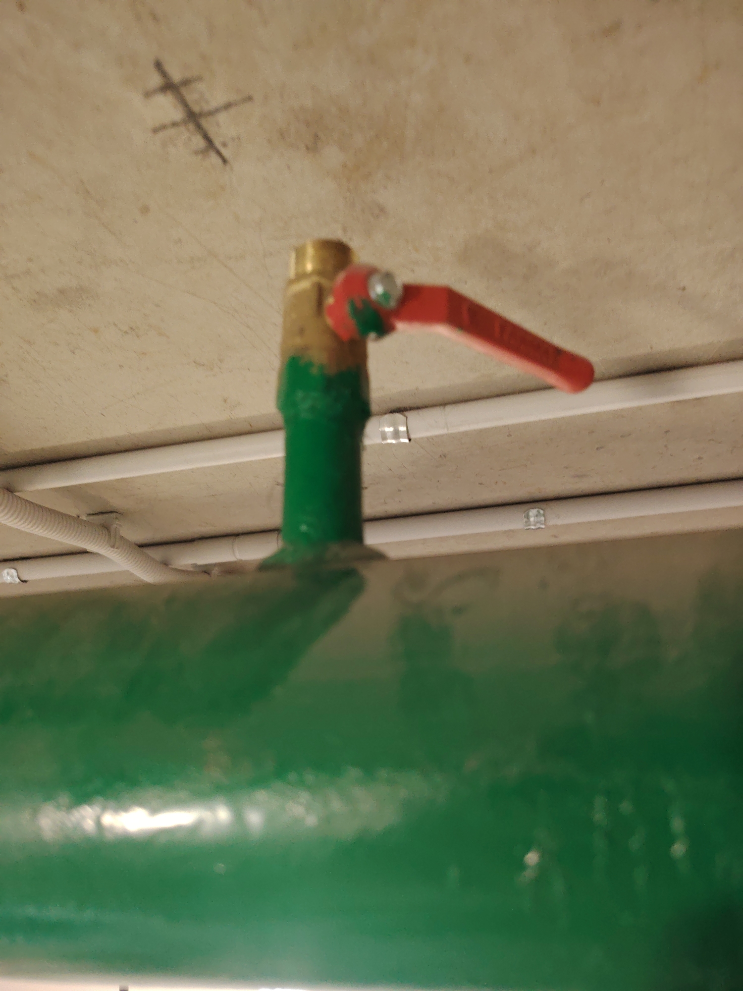An overhead view captures a section of industrial piping against a concrete ceiling. A green pipe dominates the foreground, reflecting light and obscuring much of the lower half of the image. Above the green pipe, a smaller vertical pipe extends upwards, painted green at its base and transitioning to a brass fitting. On top of the brass fitting is a red ball valve with a silver lever, positioned horizontally. The concrete ceiling above is textured and shows a faint marking resembling a stylized "H" or a cross. Several white conduits, secured by metallic clips, are visible attached to the ceiling, running parallel to each other and the large green pipe. The overall scene suggests an indoor, possibly utilitarian, environment such as a utility room or industrial space.