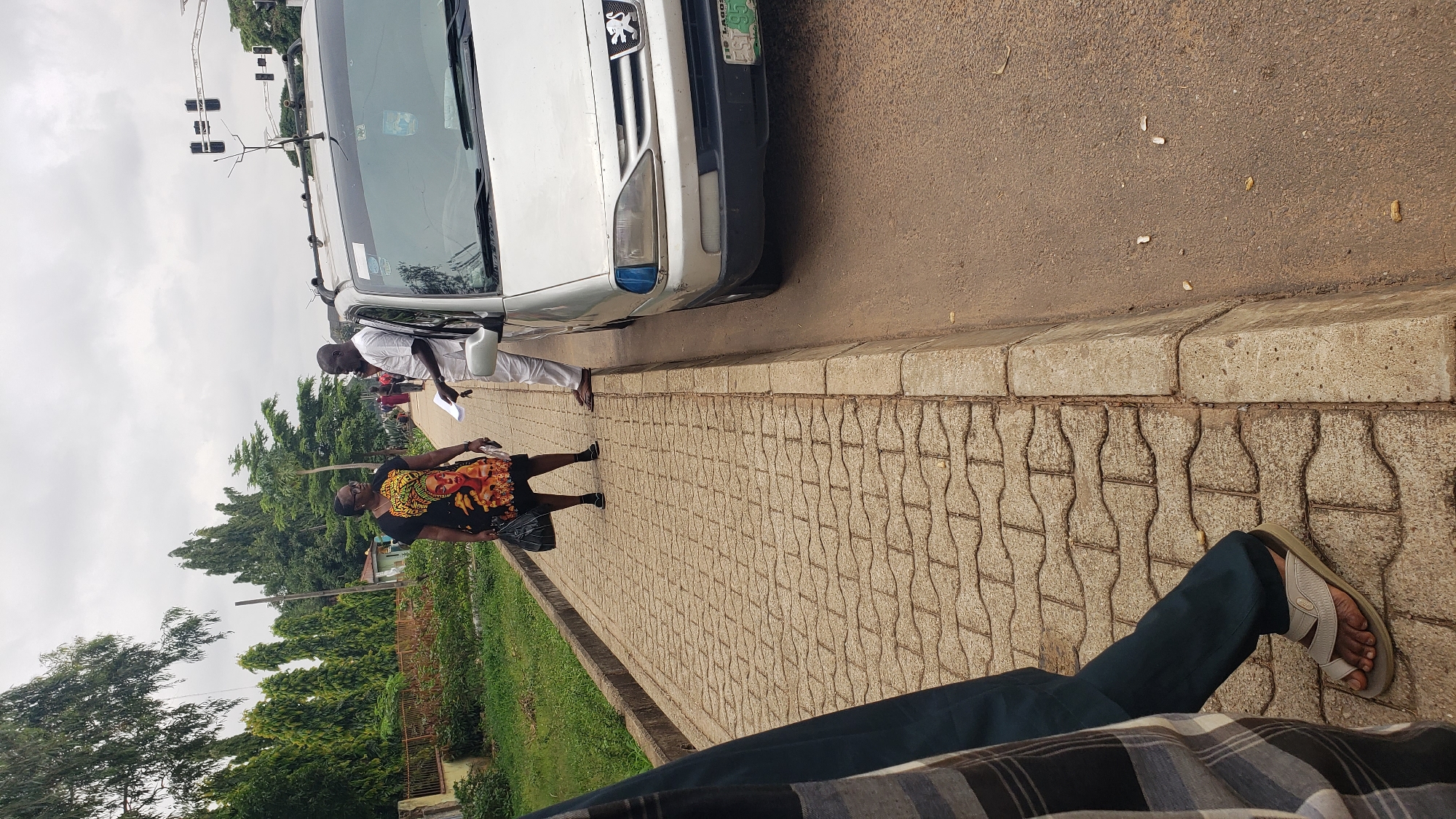 In Unguwar Shanu, Nigeria, on an overcast day, a silver sedan is parked on the right side of a road, its front passenger door ajar. A man in a white tunic and trousers stands next to the open door, facing a woman who is also standing. The woman wears a black dress with a vibrant, graphic print of a woman's face and upper body. She carries a dark bag in her left hand. Both individuals appear to be engaged in conversation. The sidewalk, paved with light brown interlocking bricks, runs parallel to the road. Beyond the sidewalk, a line of well-maintained, conical trees borders a grassy area. In the background, traffic lights are visible above the trees. The foreground shows the lower legs and feet of another individual wearing dark trousers and sandals, walking on the sidewalk. The scene is captured from a slightly elevated perspective, looking down and across the sidewalk towards the vehicle and the two conversing individuals.