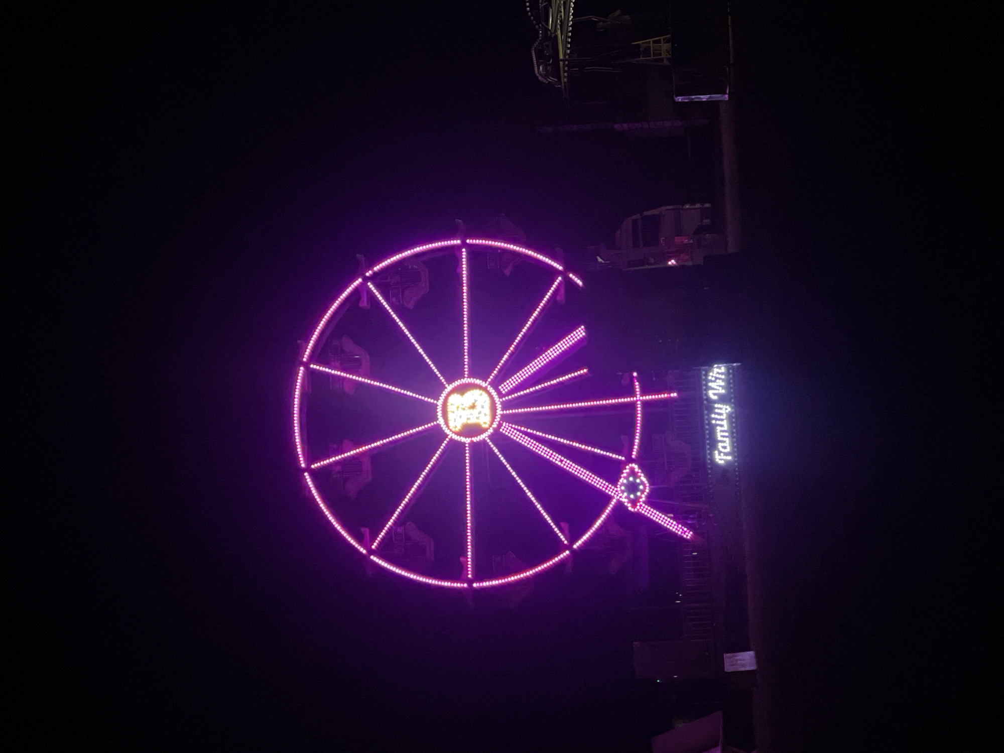 The image depicts a Ferris wheel at night, brightly lit with purple neon lights. The Ferris wheel is the main subject, with cabins visible along its circumference. To the right of the Ferris wheel is a structure with the words "Family Wheel" displayed vertically in illuminated letters. Other structures are visible in the upper right of the image. The background is mostly dark, suggesting a night scene. The location is not specified, and there is no indication of it being in Kazanlak, Bulgaria.