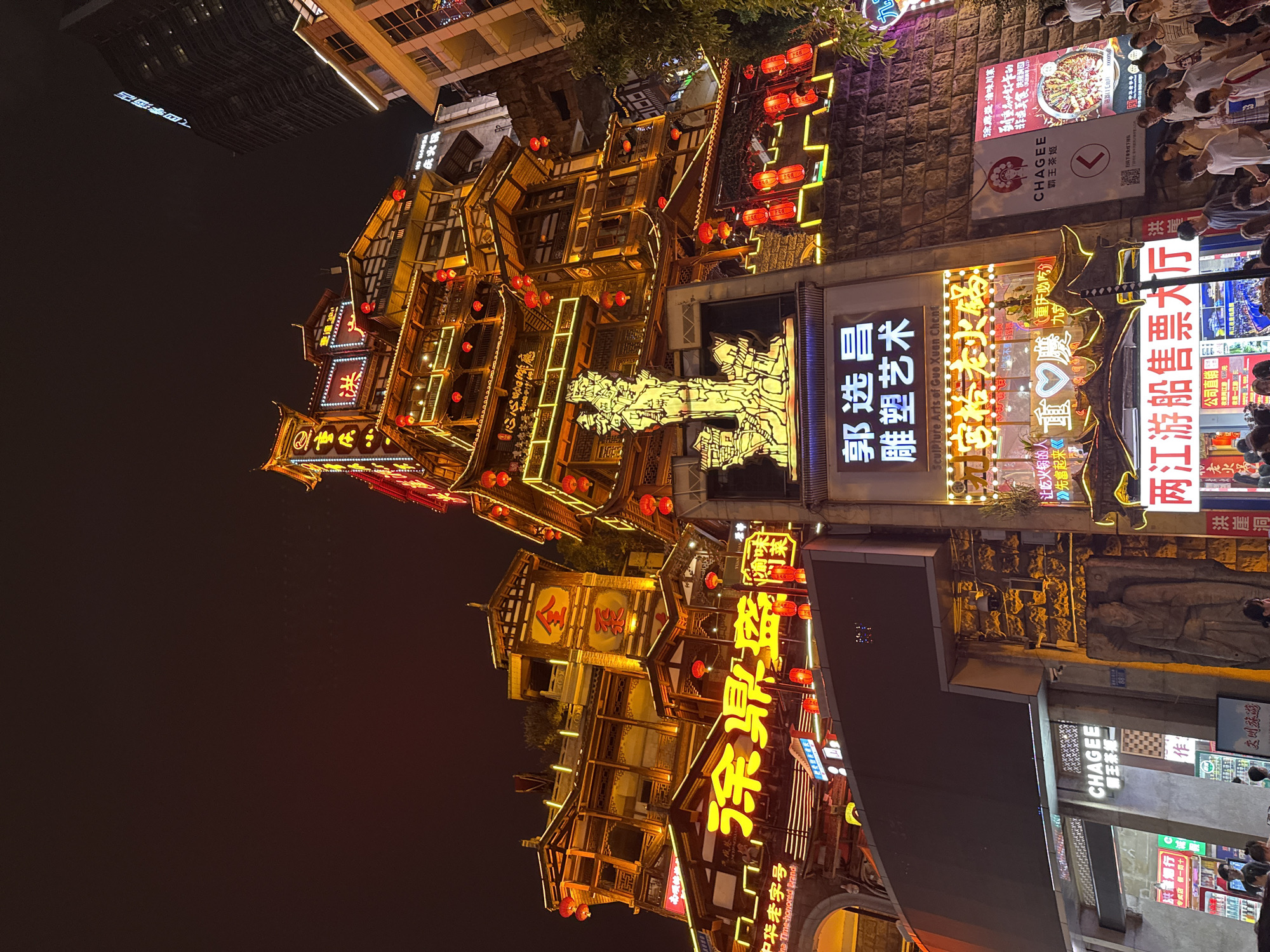 The image depicts a nighttime scene in Yuzhong District, China, showcasing a multi-story traditional Chinese building illuminated by numerous lights and red lanterns. The building features intricate wooden architecture with multiple balconies and decorative elements. Prominent neon signs in Chinese characters are visible on the facade, advertising businesses and attractions. A large digital screen is mounted on the building, currently dark. In the foreground, a crowd of people is gathered, some looking up at the building, others walking. The environment is urban, with other buildings and structures visible in the background, including a tall, modern skyscraper to the right. The overall atmosphere is vibrant and bustling, characteristic of a lively commercial district at night.