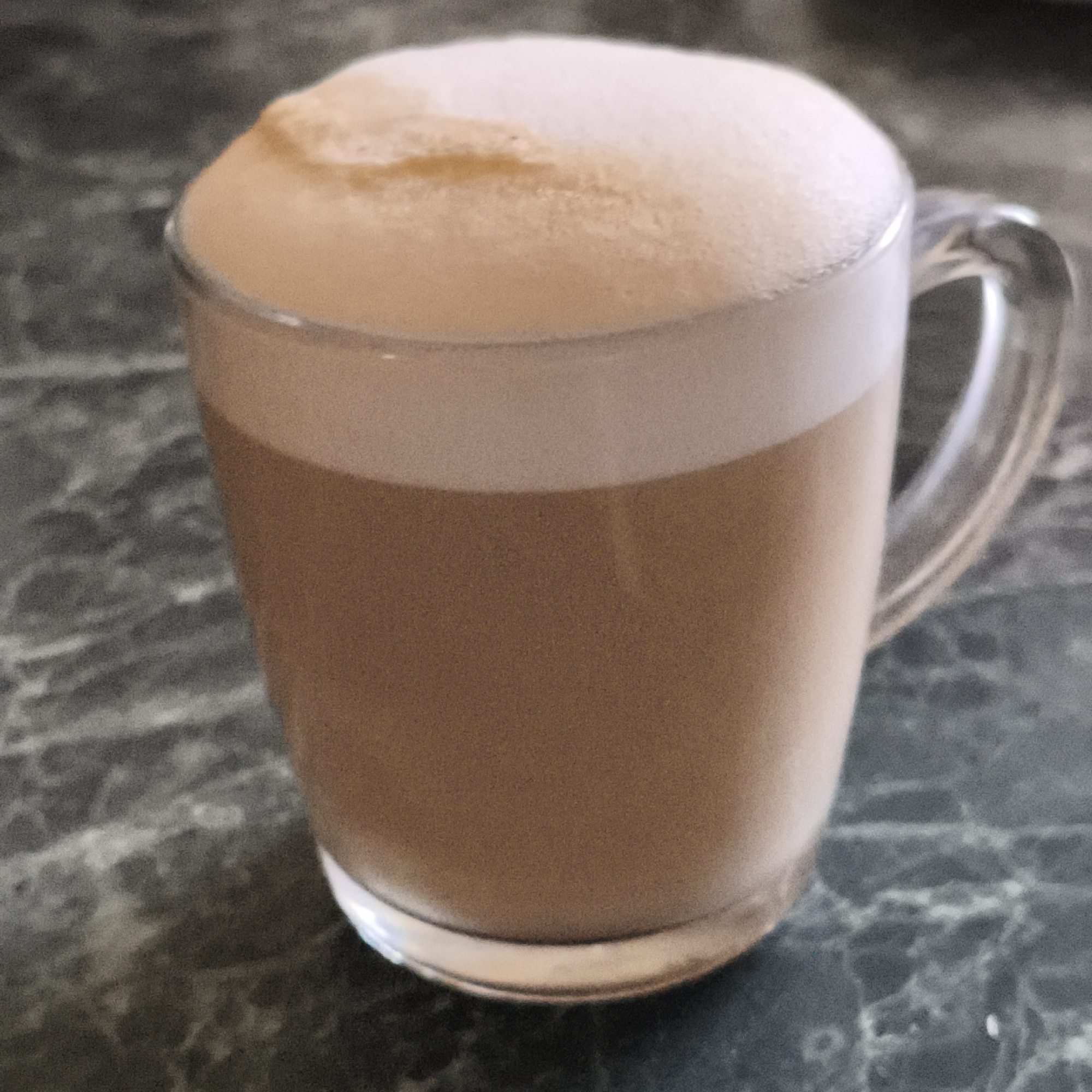 A glass mug filled with a frothy beverage, possibly a latte or cappuccino, sits on a green-gray marble-like surface. The mug is clear with a handle and the beverage is light brown with a thick layer of off-white foam. The scene is indoors. The location is Unknown, Unknown.