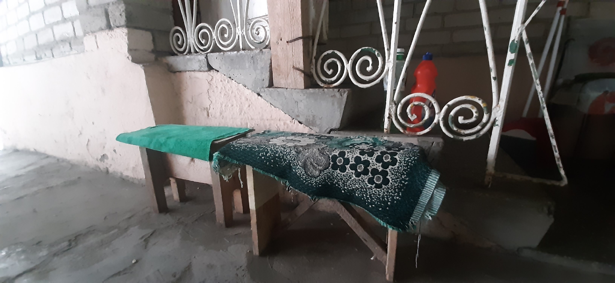 The image depicts an outdoor scene in Yessentukskaya, Russia. In the foreground, two wooden benches are visible. The left bench is covered with a bright green cloth, while the right bench is adorned with a patterned dark green textile featuring floral designs. Behind the benches, a concrete staircase with decorative white metal railings leads upwards. To the right of the staircase, a red plastic bottle with a blue cap is placed near the railings. The ground appears to be concrete or dirt. The overall environment suggests a simple, possibly aged, outdoor residential or communal area. There are no people present in the image.