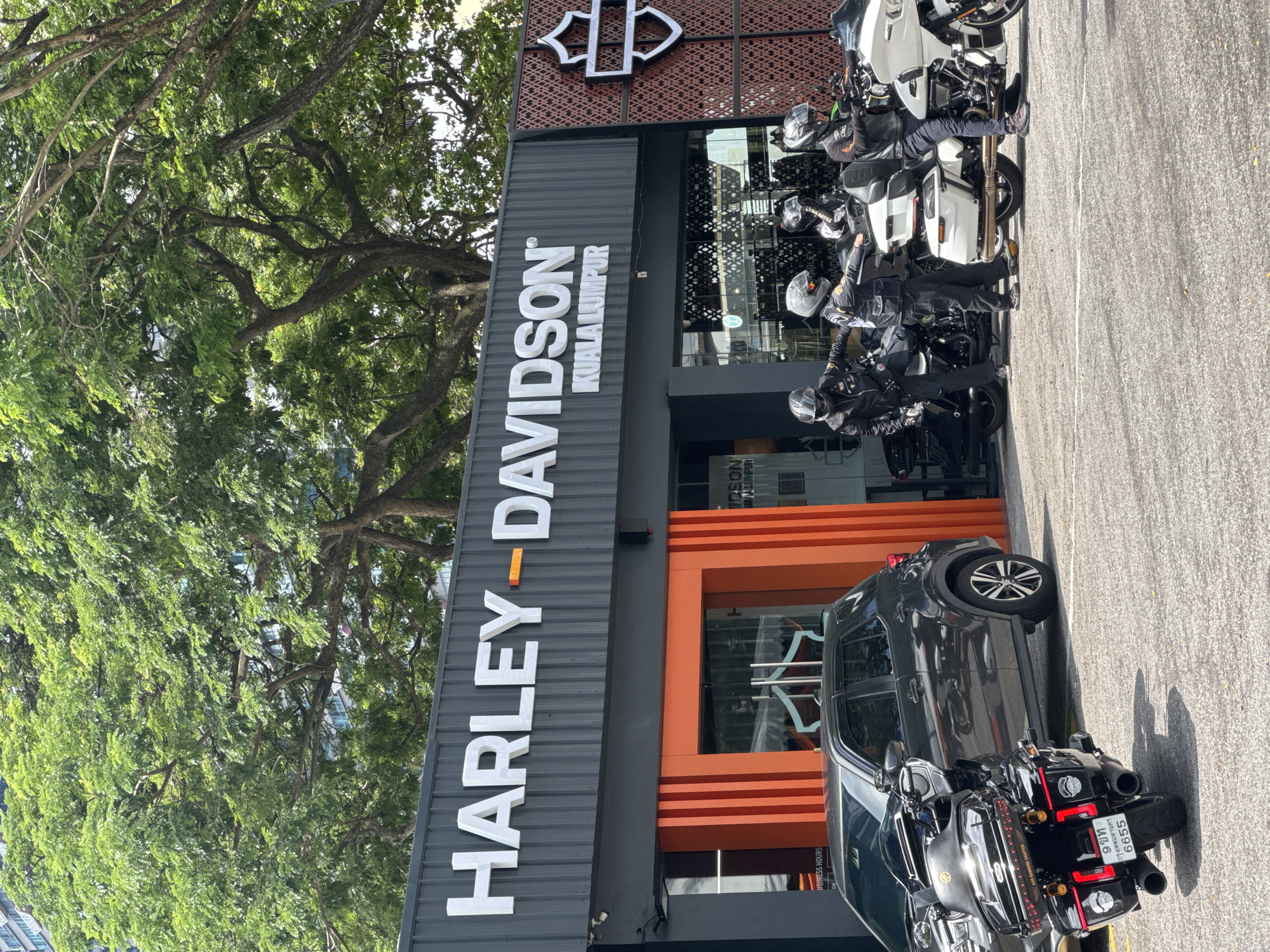 The image depicts the Harley-Davidson dealership in Kuala Lumpur, Malaysia. The dealership's black facade features the brand name in white lettering with an orange highlight. An adjacent orange-framed window with orange horizontal bars is visible. Parked in front are several Harley-Davidson motorcycles, partially obscured by a dark-colored SUV. The motorcycles have uniformed individuals, potentially officers, sitting on them. A large tree with dense green foliage is to the left of the building. A white motorcycle is parked near the right of the storefront.