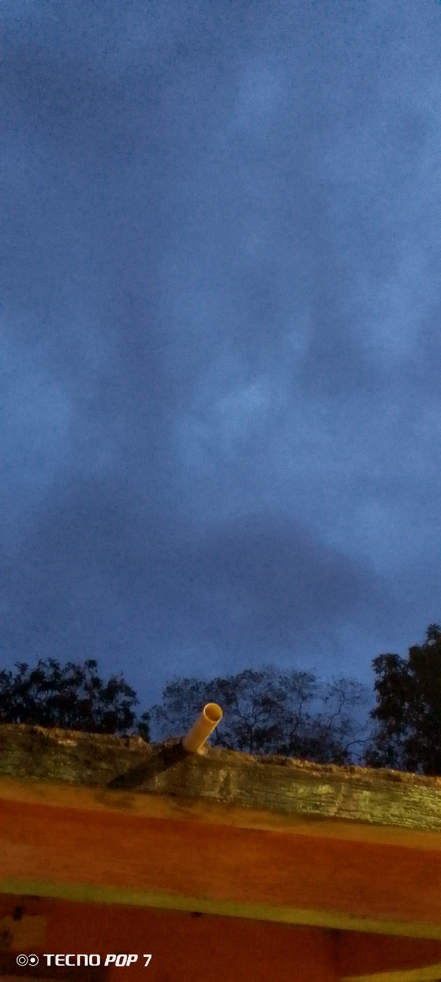 The image depicts a cloudy, dark blue sky over a line of silhouetted trees. In the foreground, a bright yellow pipe juts out from a textured surface, casting a shadow. The lower portion of the image is dominated by an orange and yellow structure. A watermark in the bottom left corner reads "TECNO POP 7." The scene appears to be an outdoor, possibly urban, setting in Altagracia, Venezuela, under twilight or overcast conditions. There are no people or discernible actions in the image.
