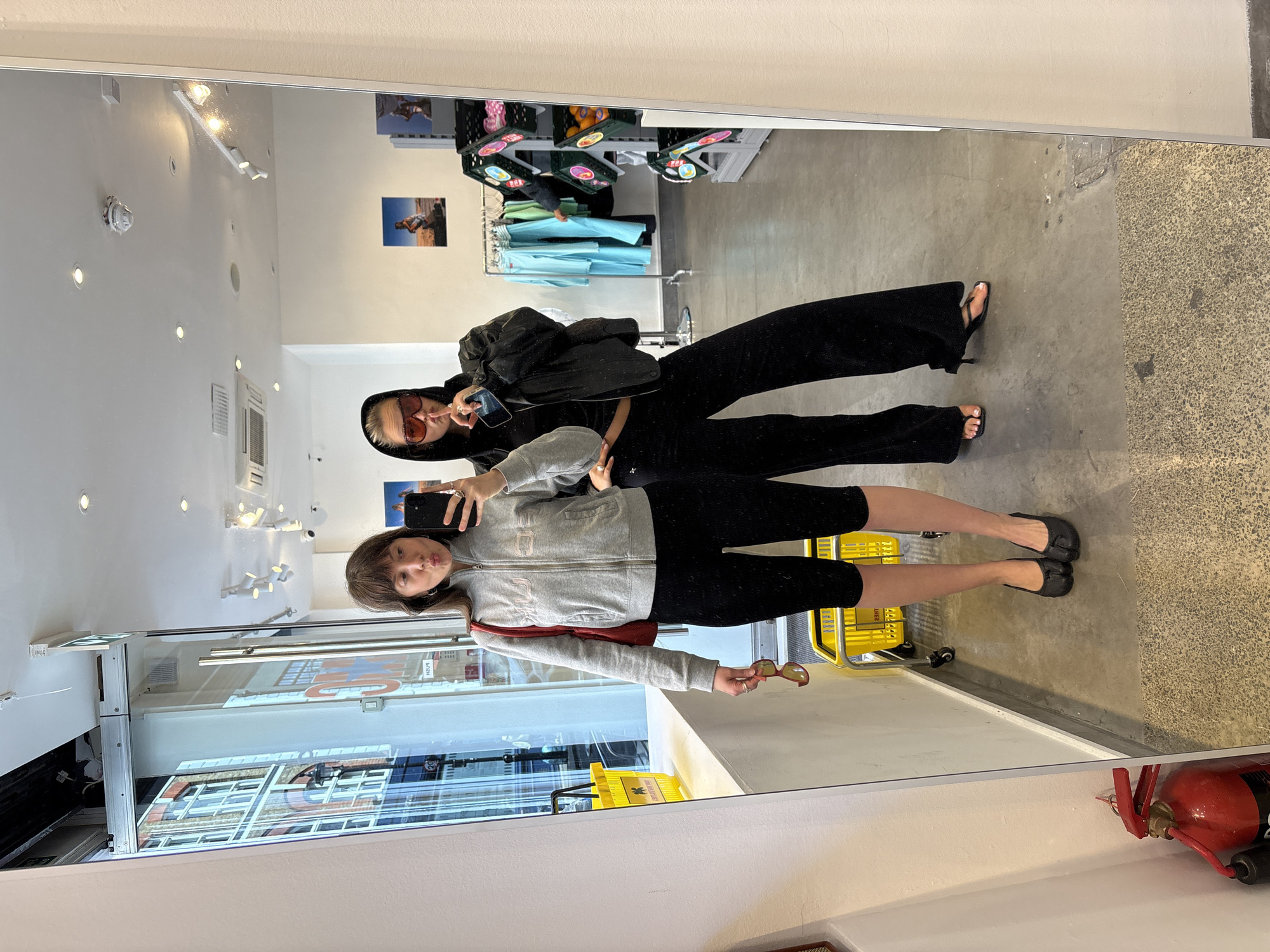 Two women are standing in front of a mirror inside a retail store, taking a selfie. The mirror reflects the interior of the store, showcasing clothing racks, display shelves, and other merchandise. The store's entrance, with glass doors and windows, is also visible in the reflection, revealing an urban street scene outside. The exterior view suggests the store is located in London, United Kingdom, based on the architectural style of the buildings across the street. One woman is wearing a grey zip-up jacket, black leggings, and black flats. She is holding a phone in one hand and sunglasses in the other. The other woman is wearing a black hooded jacket, black pants, and sandals. She is holding a phone in one hand and making a face. The overall scene depicts a casual moment captured in a retail environment.