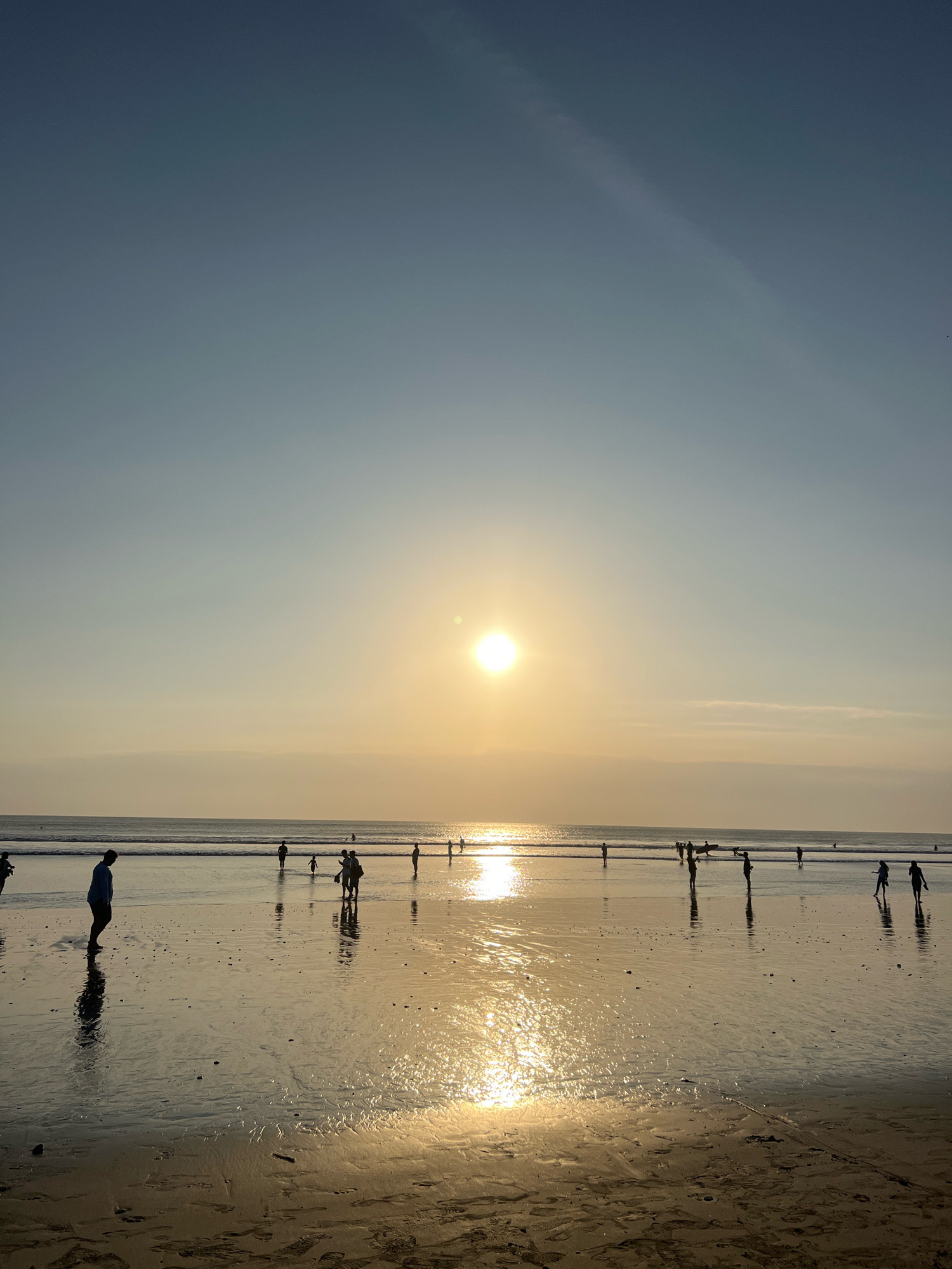 The image captures a sunset scene on Kuta Beach, Bali, Indonesia. The sun, a bright orb, is positioned in the upper center of the frame, casting a warm, golden glow across the sky and reflecting intensely on the wet sand. The sky transitions from a pale blue at the top to a soft yellow and orange hue near the horizon. Gentle waves roll onto the shore, creating a wet, reflective surface on the beach.
Several individuals are present on the beach, silhouetted against the bright sky and the sun's reflection. In the foreground, a person in a blue shirt stands with their back to the camera, casting a distinct shadow. Further down the beach, a group of people are scattered, some walking, some standing, and one appears to be holding a surfboard. Their figures are dark against the luminous background. The sand shows evidence of footprints and the rippling texture left by receding water. The overall atmosphere is tranquil, with the natural beauty of the sunset dominating the scene.