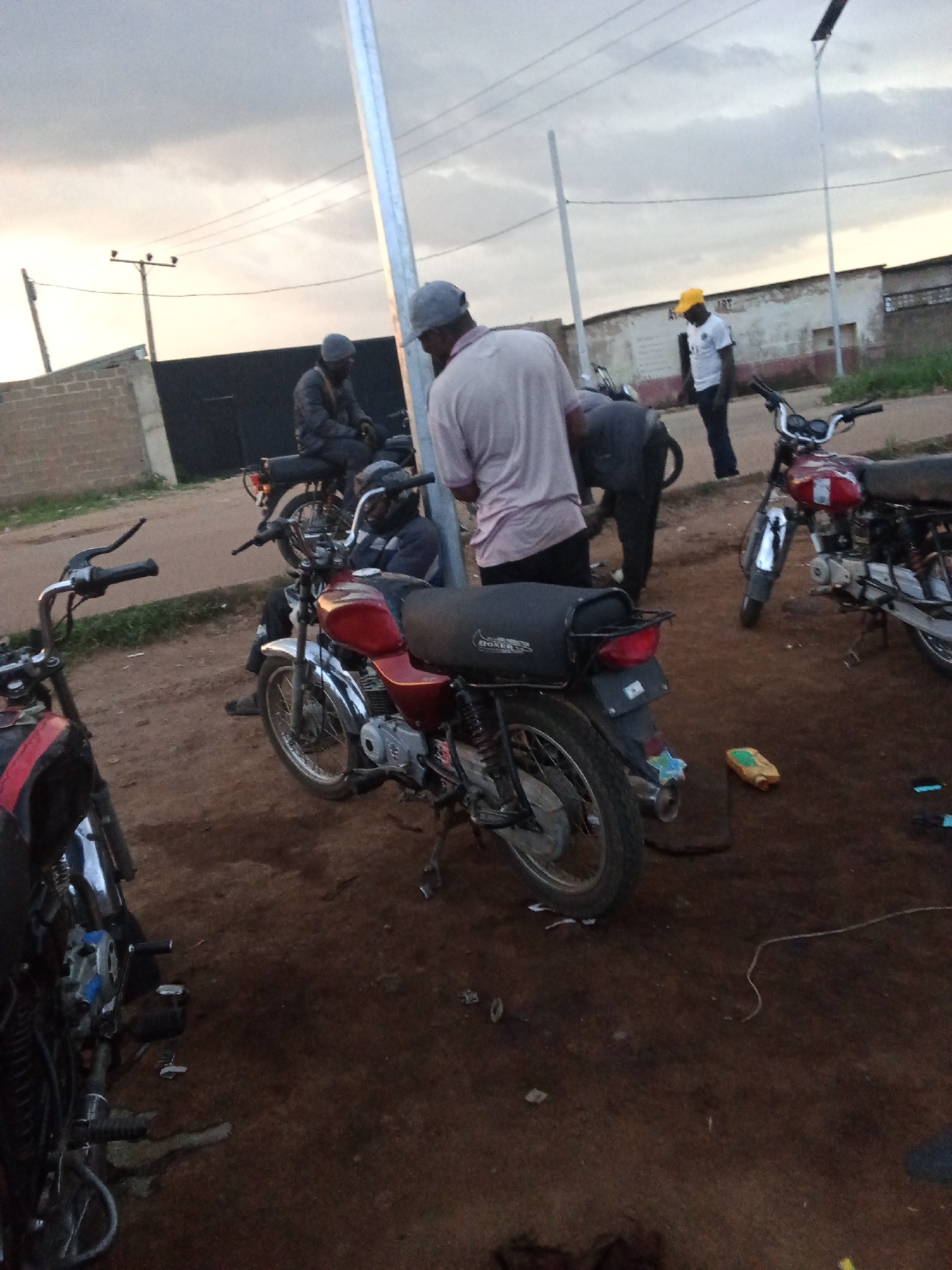 In Barakin Lerawi, Nigeria, on a dusty, unpaved road beneath an overcast sky, a group of individuals are gathered around several motorcycles. The scene appears to be an informal gathering or possibly a repair area.
In the foreground, a red and black motorcycle is positioned at an angle. Its front wheel is visible, and the handlebars are turned slightly to the left. To the left of this motorcycle, the rear of another motorcycle, predominantly black with some red accents, is visible.
Behind the red and black motorcycle, a man wearing a light pink shirt and a grey cap is standing, facing away from the camera. To his left, another person in a dark jacket and grey cap is seated on a motorcycle, also facing away. Further back, to the right of the pink-shirted man, a person in a white t-shirt and a bright yellow cap is standing and looking down. To the far right, a red motorcycle is parked, with its front wheel and handlebars visible.
The ground is covered in loose, dark brown dirt and scattered debris, including what appears to be a yellow and green plastic bottle. In the background, a brick wall and a dark, solid fence line the area. Power lines and poles are visible against the cloudy sky, indicating an urban or semi-urban setting. The overall impression is one of everyday life, with people and their vehicles in a common outdoor space.
