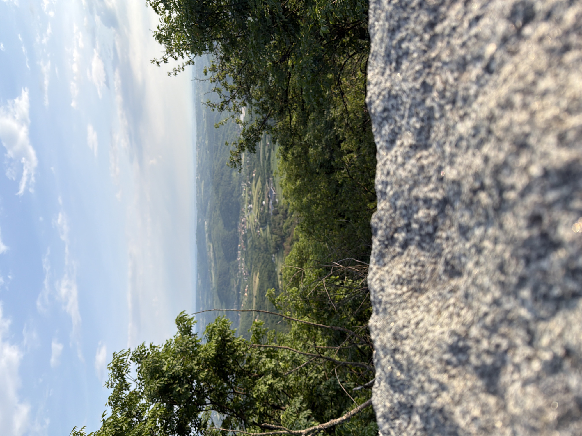 The image shows a view from a rocky outcrop on a hill or mountain in Vozdovac Urban Municipality, Serbia. The foreground includes a large, light-colored rock formation, and branches with green leaves. In the distance, a valley with trees and buildings is visible. The sky is blue with some clouds.