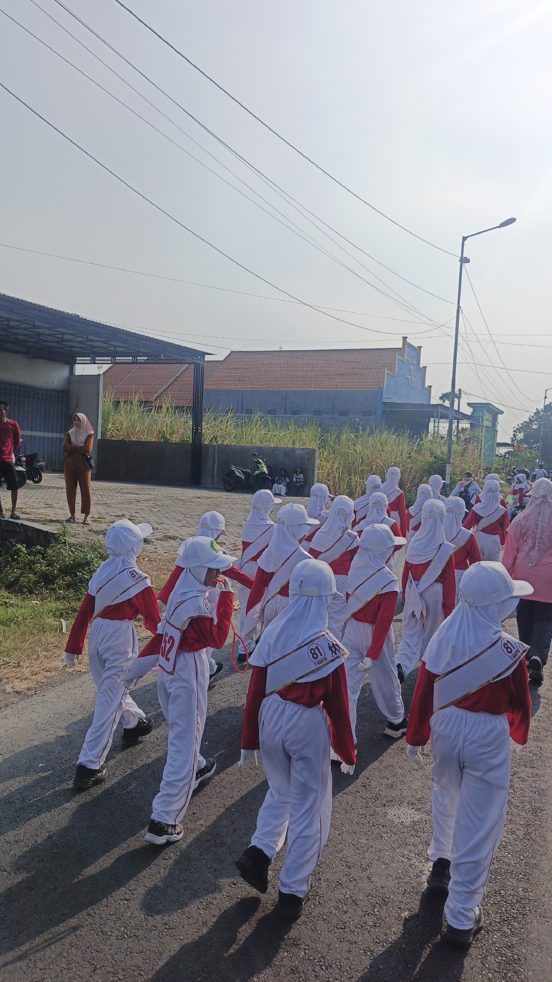 In Puri, Indonesia, a group of children, likely students from a school, are participating in a parade. They are dressed in matching uniforms consisting of white pants, white headscarves, and red tops with white sashes. The sashes are adorned with golden trim and some have the number "81" printed on them, possibly indicating a school or group number. The children are walking in formation on an asphalt road, casting long shadows on the ground due to the bright sunlight. The background shows a street lined with low buildings, some with tiled roofs, and an area with tall dry grass. A street lamp is visible on the right side of the image, and power lines crisscross the sky. In the distance, a few adults and motorcycles are also present, suggesting the presence of observers or support staff for the parade.